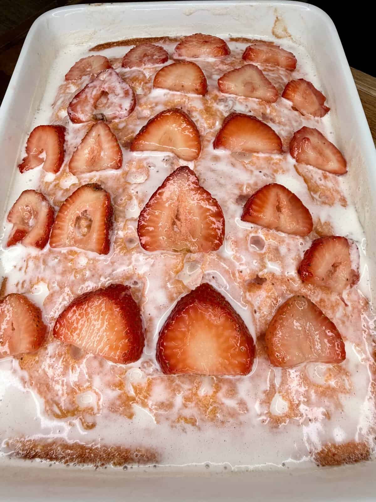 A close-up of a baked dessert in a white dish, topped with sliced fresh strawberries and a drizzle of glaze or icing. The surface appears golden brown with a shiny, creamy coating.