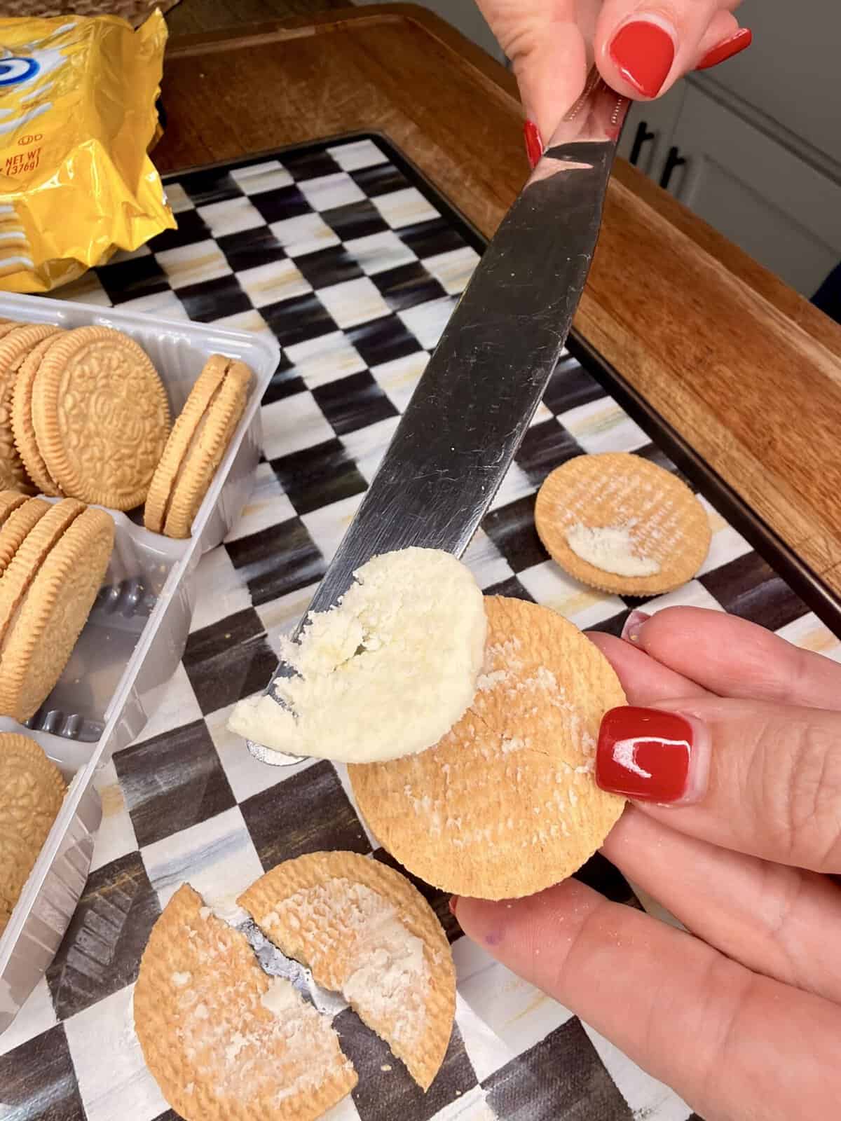 A person with red nail polish uses a knife to scrape cream filling from a golden sandwich cookie. Other cookies are nearby on a checkered surface, and one cookie is broken in half.