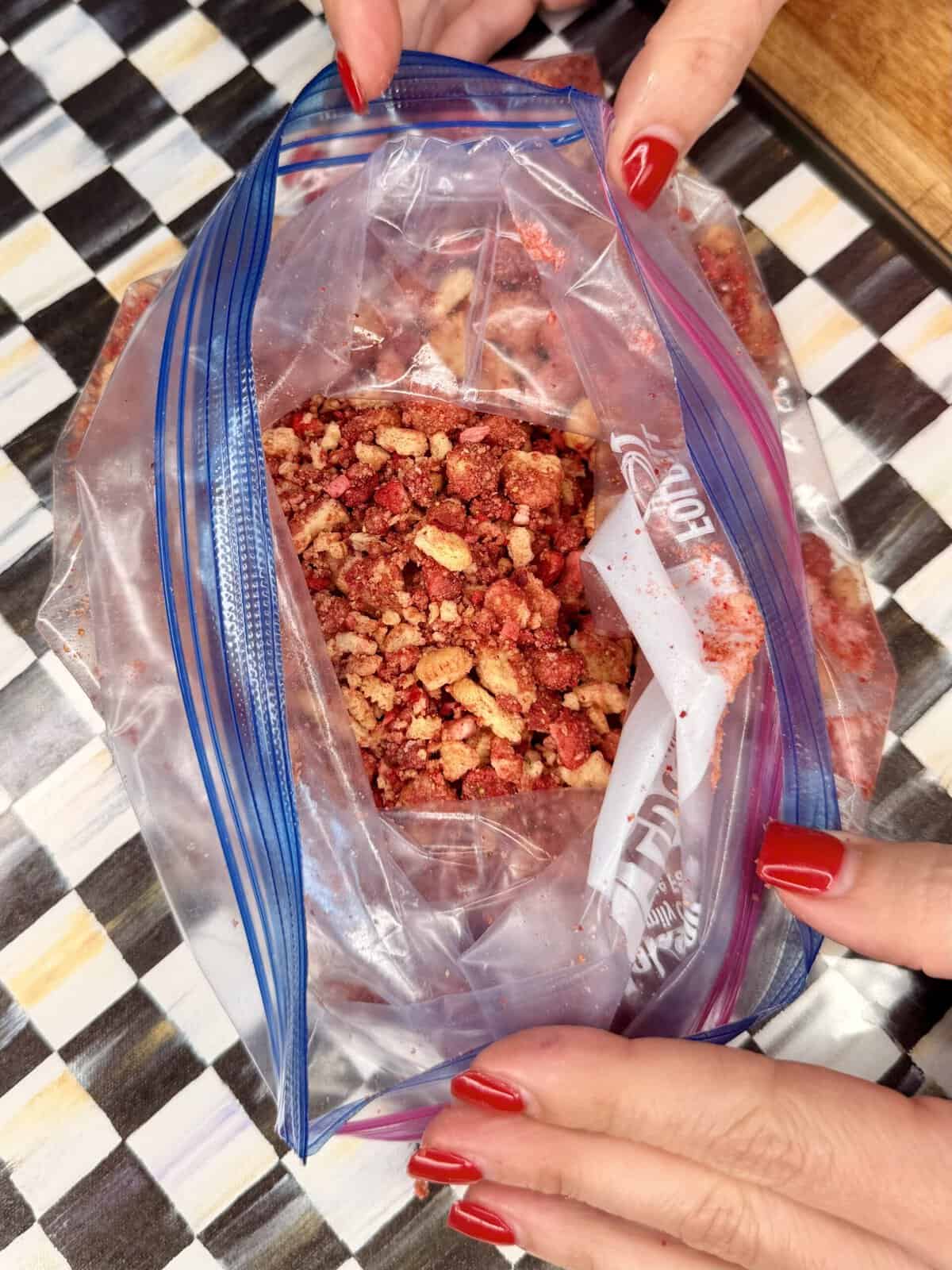 A person with red painted nails holds an open plastic ziplock bag filled with crushed red and tan-colored snacks or crumbs on a checkered surface.