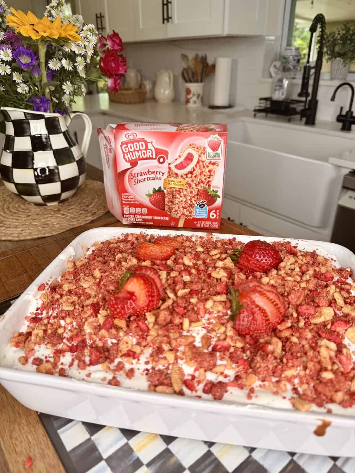 A baking dish filled with strawberry shortcake dessert topped with crumbled red and golden crumbs and fresh strawberries sits on a kitchen counter, with a Good Humor Strawberry Shortcake ice cream box in the background.