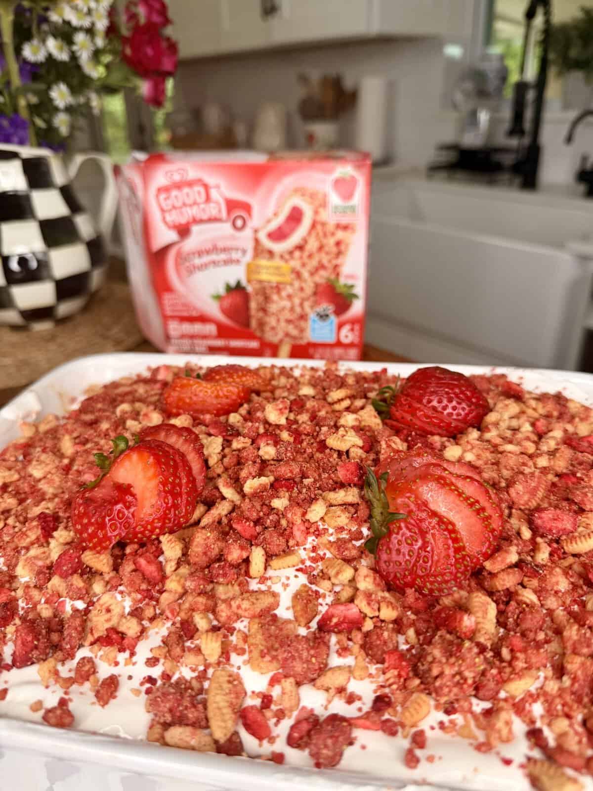 A dessert topped with crumbled strawberry shortcake bars and fresh strawberries sits on a counter, with a box of Good Humor Strawberry Shortcake bars in the background in a bright kitchen.