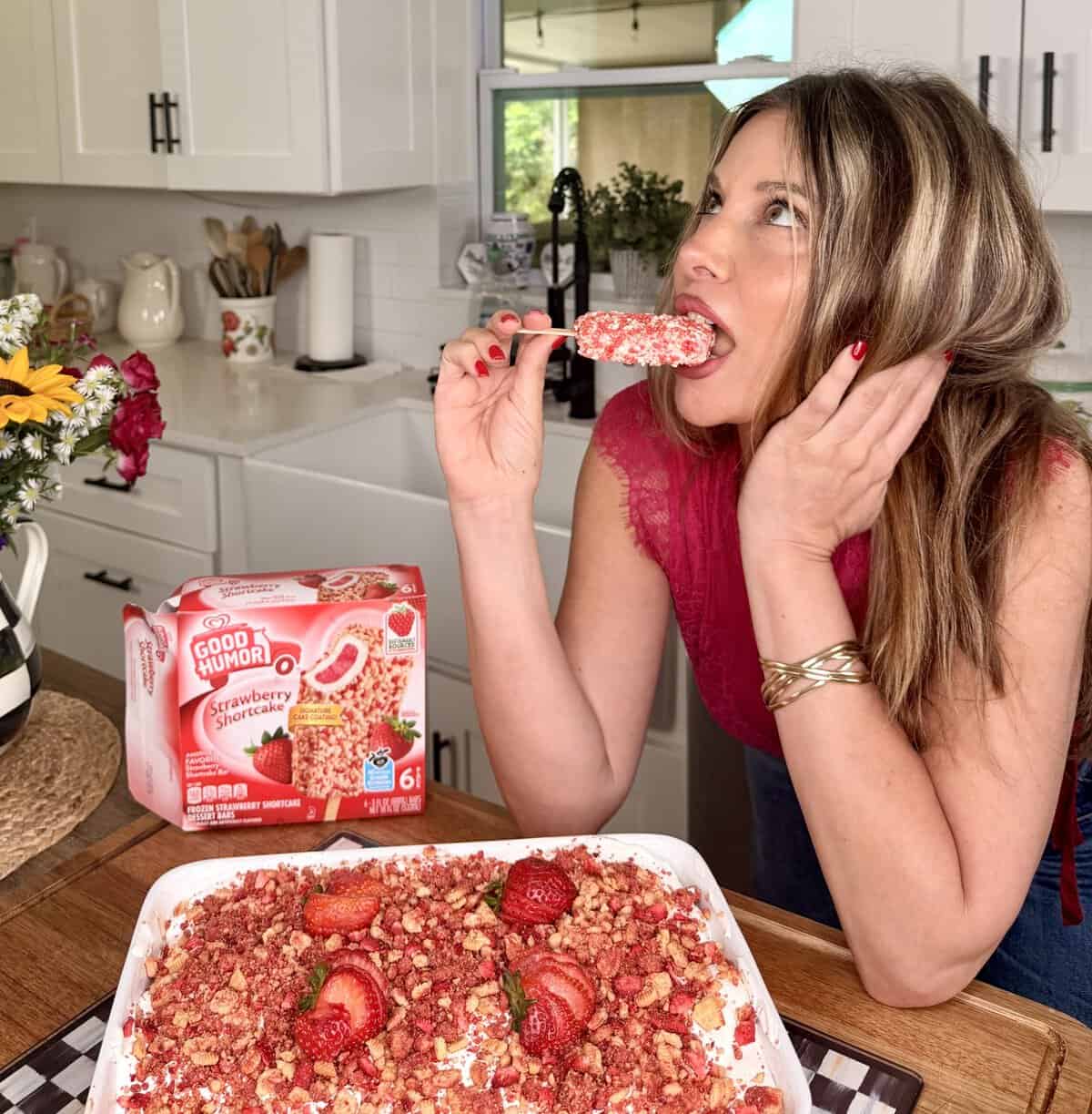 A woman in a pink top enjoys a strawberry shortcake ice cream bar in a bright kitchen, with a box of Good Humor bars and a matching strawberry dessert topped with crumbles in front of her.