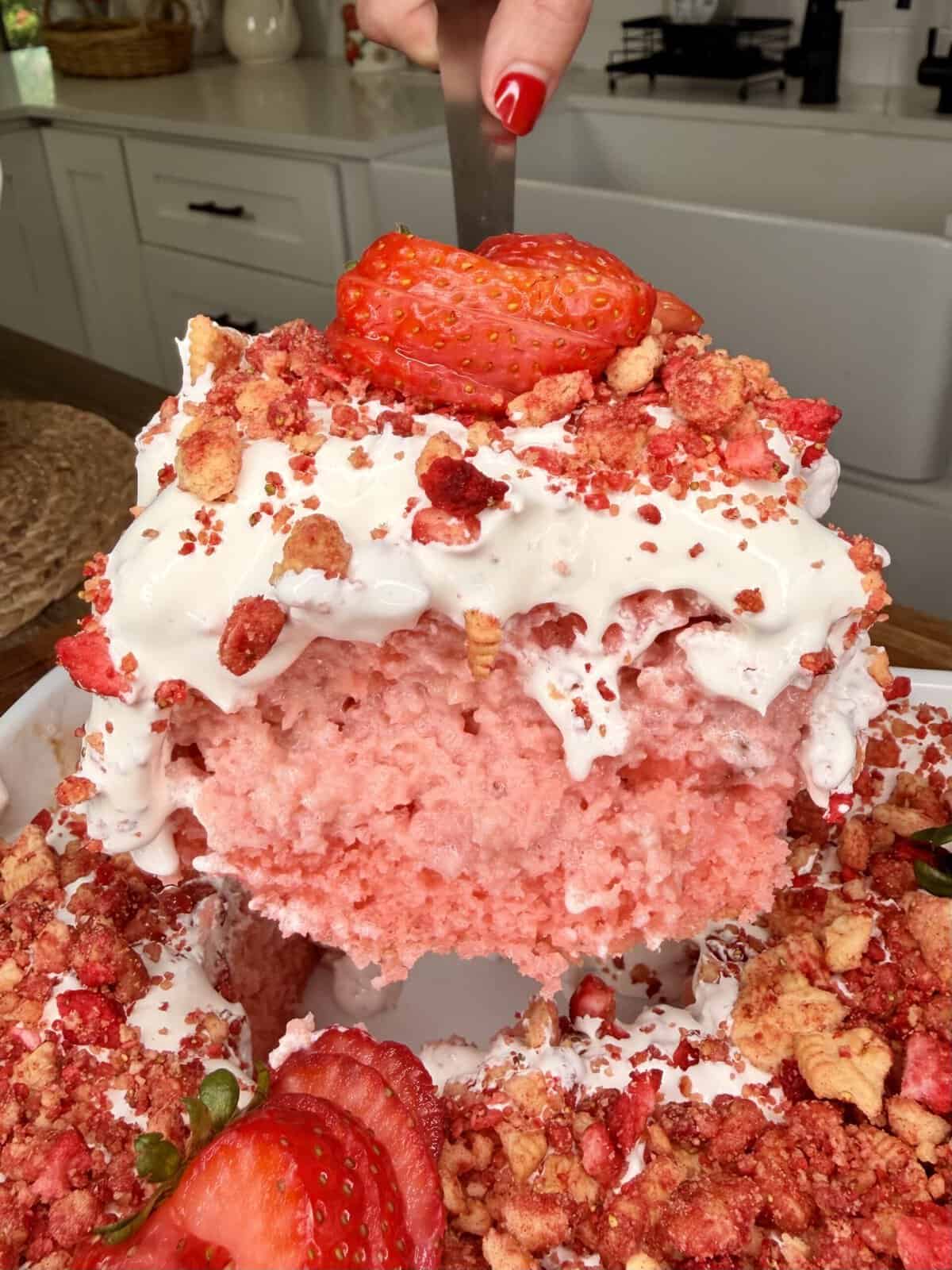 A hand lifts a large slice of pink strawberry cake topped with whipped cream, strawberry crumbles, and fresh strawberries from a bundt pan in a bright kitchen.
