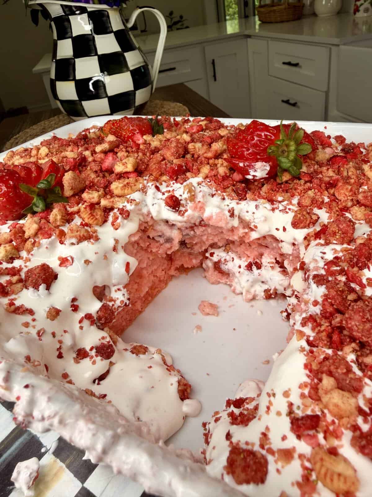 A rectangular strawberry cake topped with white frosting and red crumbly bits, garnished with fresh strawberries. A large slice is missing, and the dessert sits on a kitchen counter near a checkered pitcher.