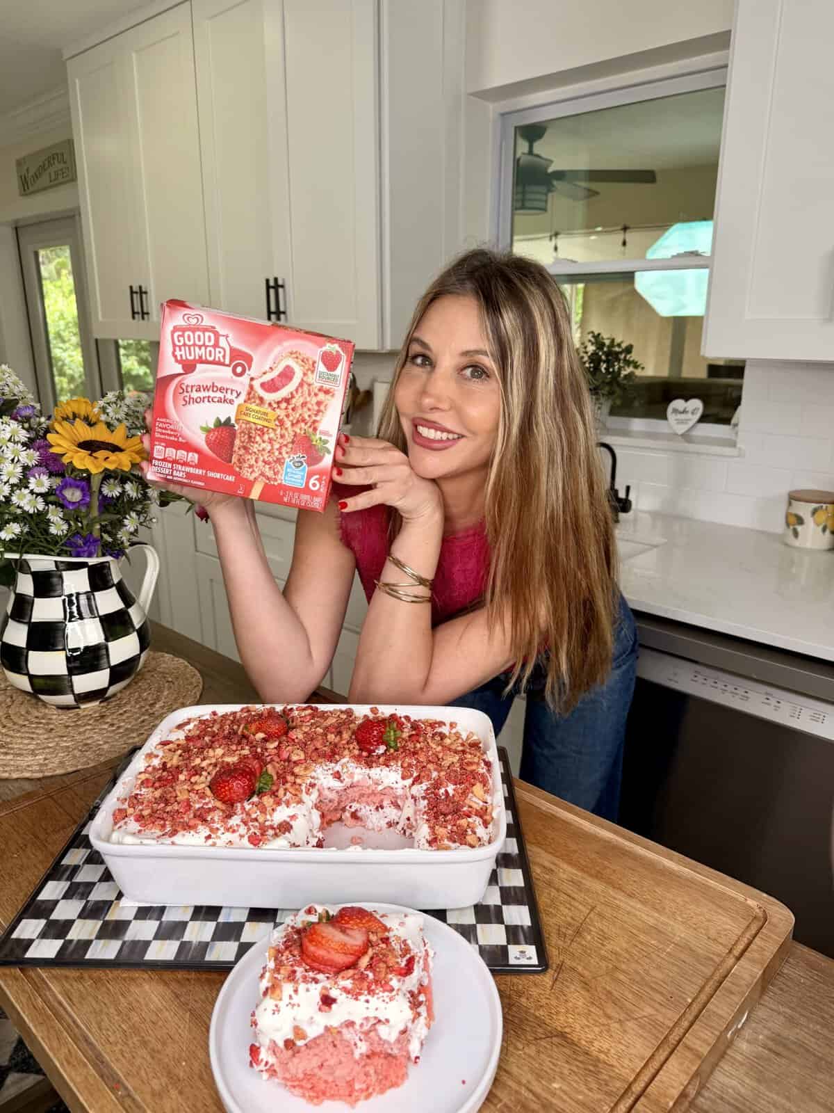 A smiling woman in a pink top holds a box of Good Humor Strawberry Shortcake ice cream bars in a kitchen, with a strawberry shortcake dessert in a dish and a plated slice in front of her. A vase of flowers is nearby.
