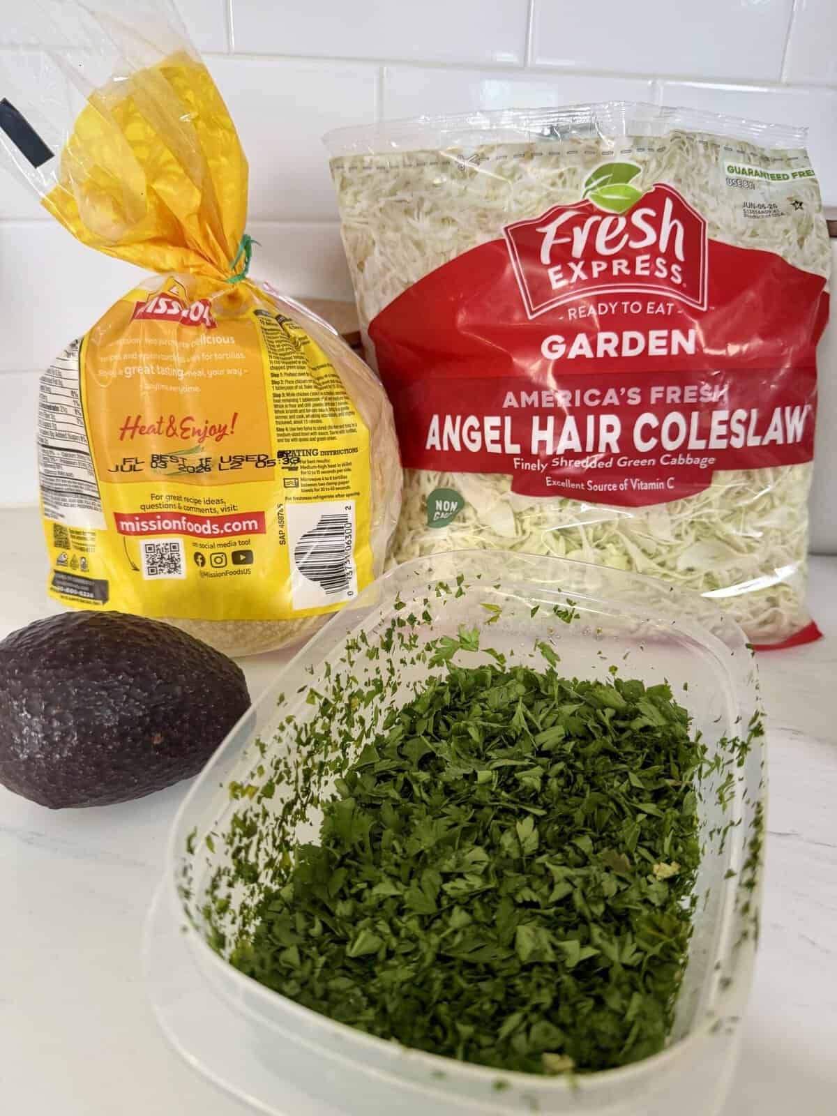 A bag of tortillas, a bag of angel hair coleslaw, a whole avocado, and a container of chopped cilantro are arranged on a white kitchen counter.