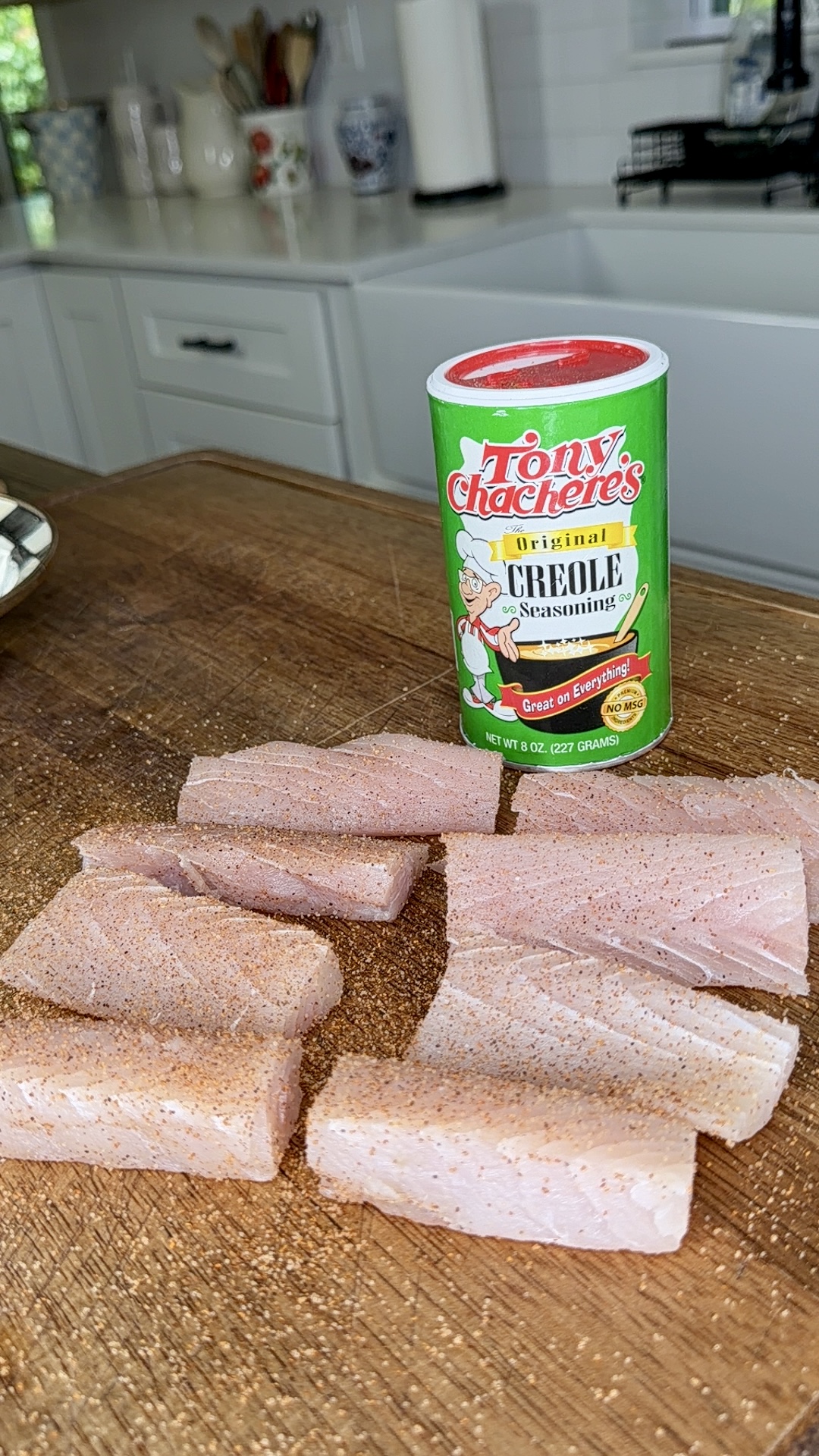 Raw fish fillets on a wooden cutting board are sprinkled with seasoning. Behind them is a canister of Tony Chachere’s Original Creole Seasoning, set in a bright, white kitchen.