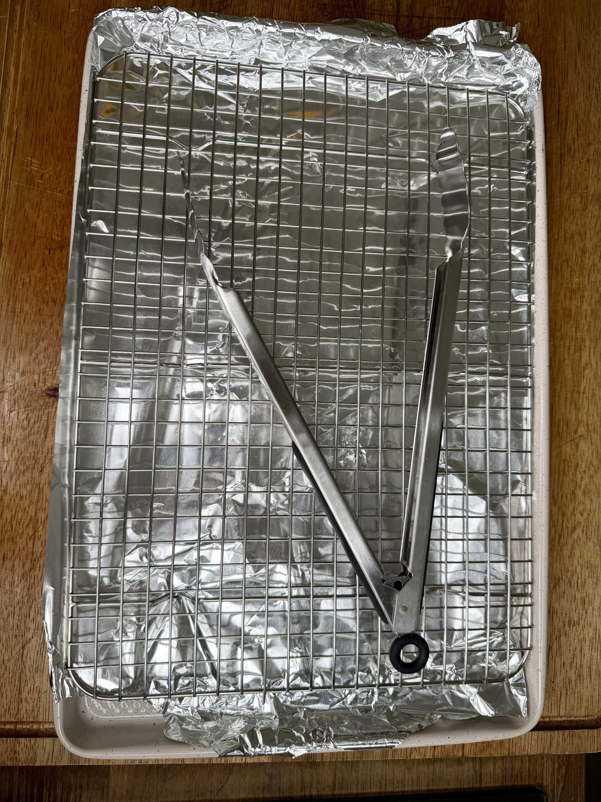 A baking sheet covered with crumpled aluminum foil, topped with a metal cooling rack. A pair of metal tongs is resting diagonally across the rack. The surface beneath appears to be wooden.