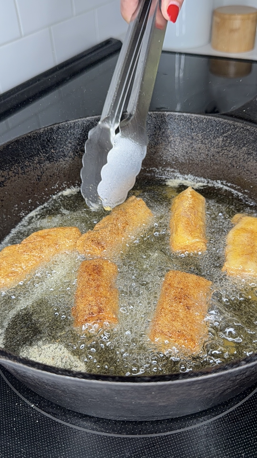 A hand using metal tongs to fry golden, breaded rolls in hot oil inside a black cast iron skillet on a stovetop.