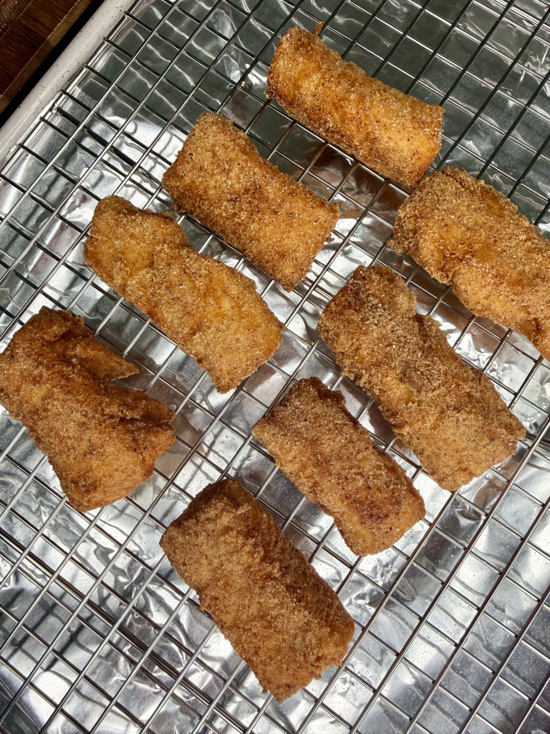 Eight golden-brown, rectangular pieces of food coated in cinnamon sugar rest on a wire cooling rack lined with aluminum foil.