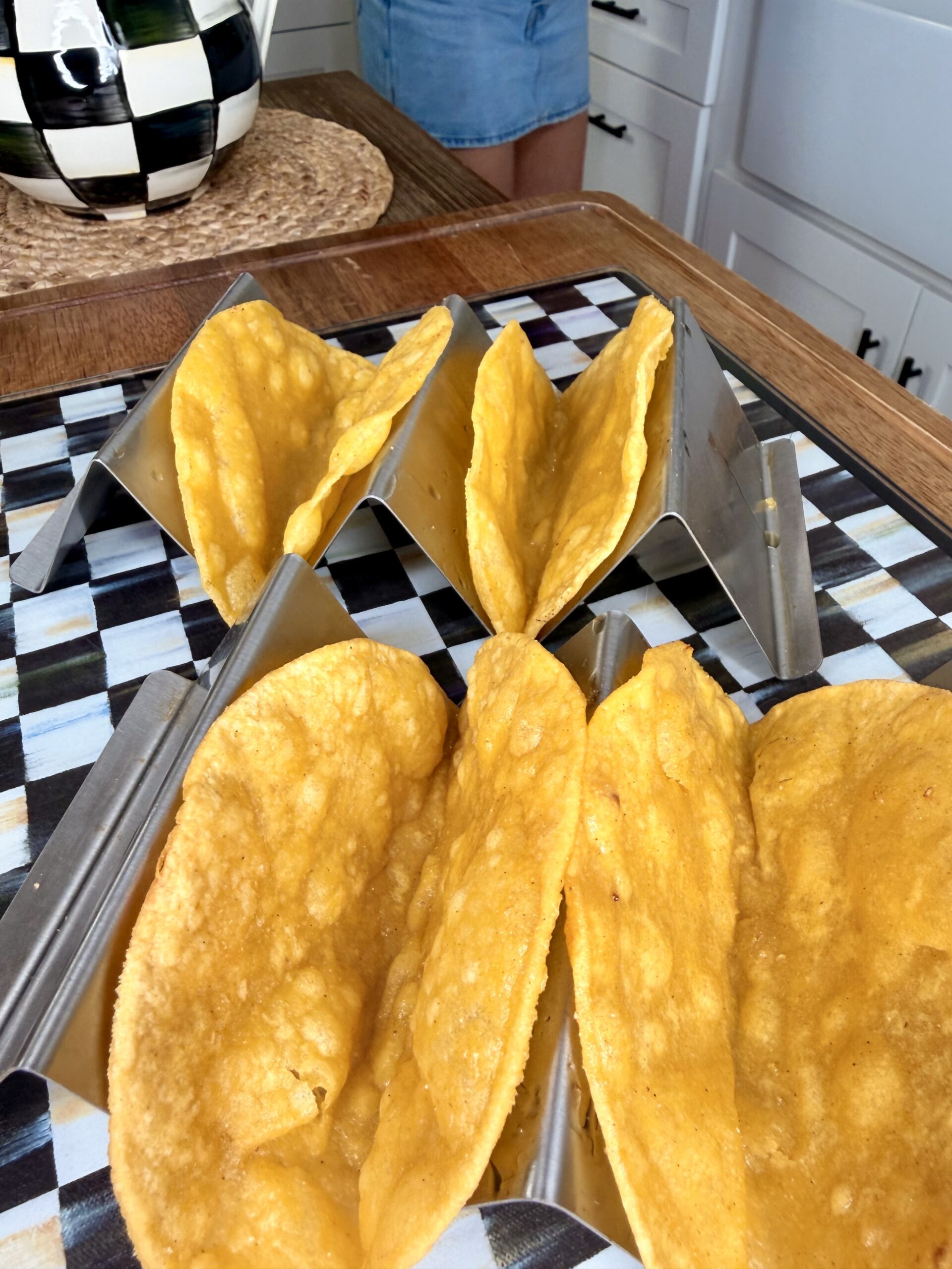 Four crunchy taco shells sit in metal taco holders on a black-and-white checkered surface, with part of a person and a kitchen background visible.