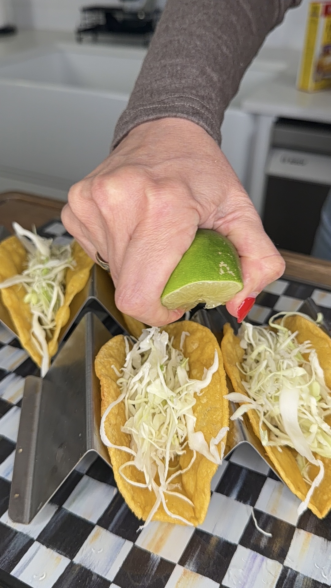 A hand squeezes a lime wedge over hard-shell tacos filled with shredded cabbage, arranged on a taco holder on a checkered surface.