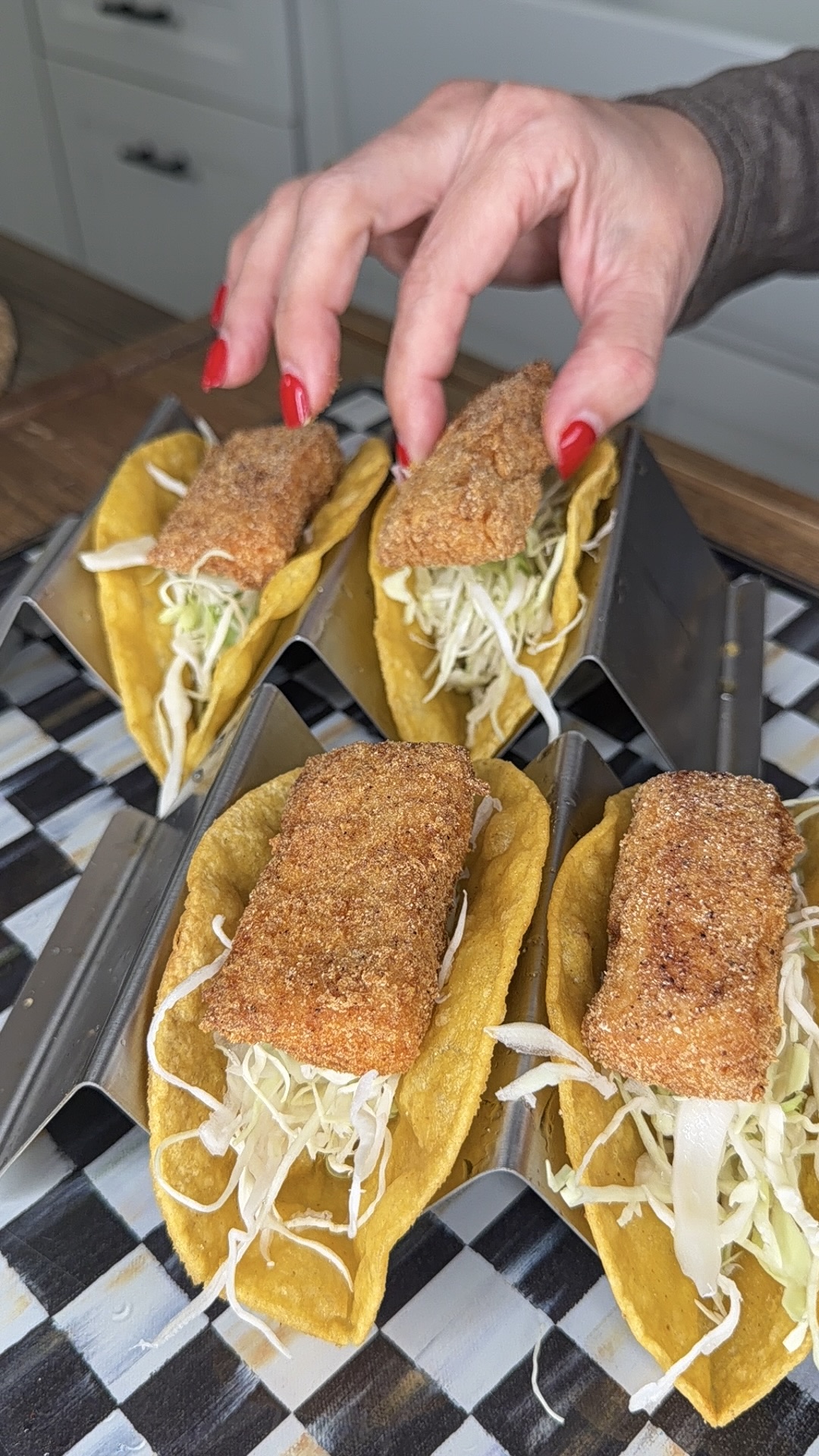 A hand with red nail polish places a piece of breaded fish on a taco filled with shredded cabbage. Three other similar fish tacos sit in metal taco holders on a checkered surface.