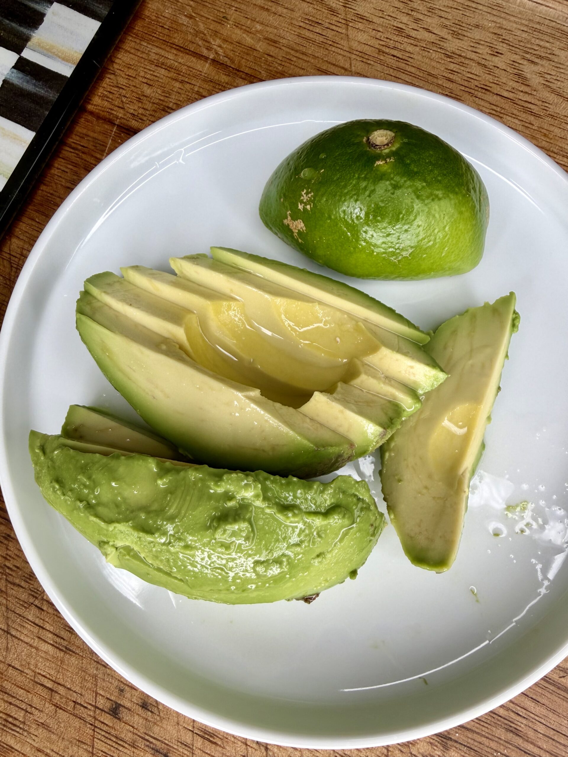 Sliced avocado drizzled with lemon juice on a white plate, next to half a lime, all placed on a wooden surface.