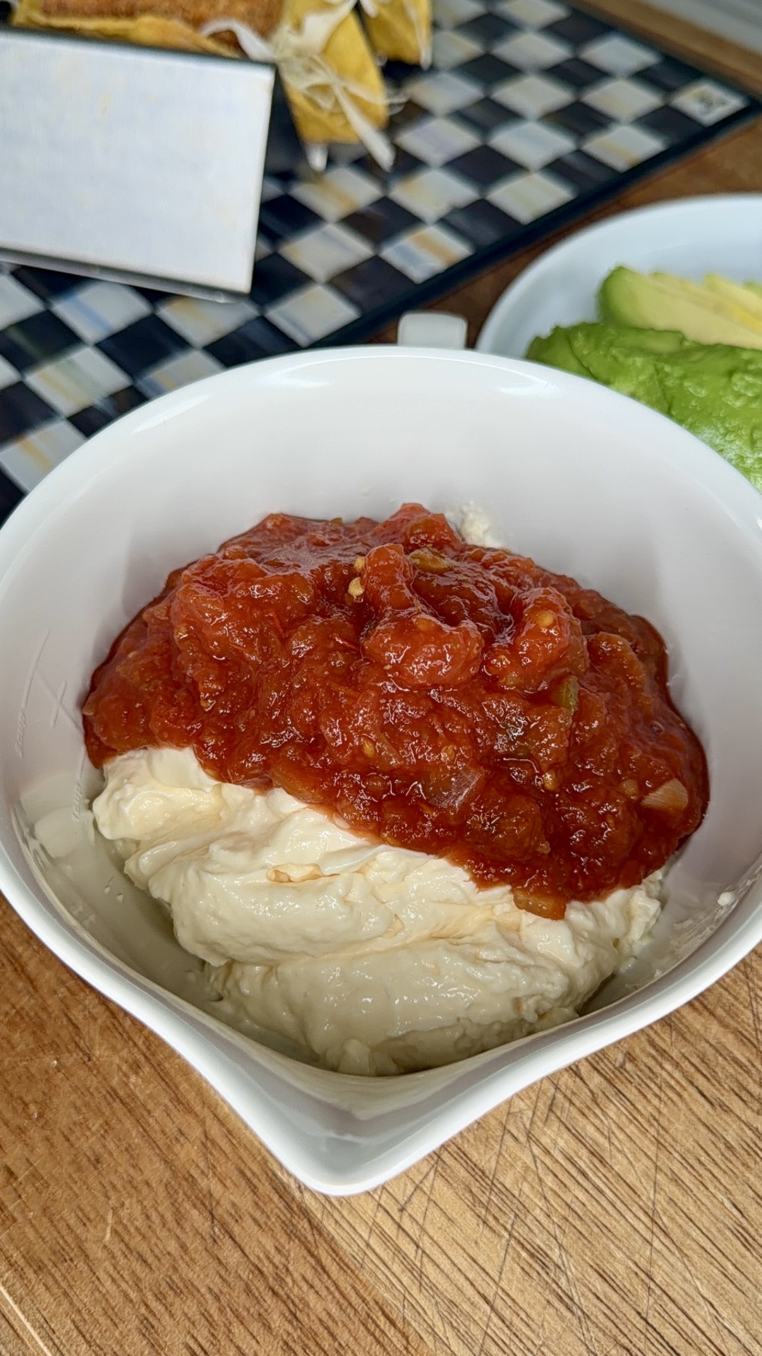 A white bowl filled with a layer of creamy white dip topped with chunky red salsa sits on a wooden surface, with sliced avocado and a checkered tray in the background.