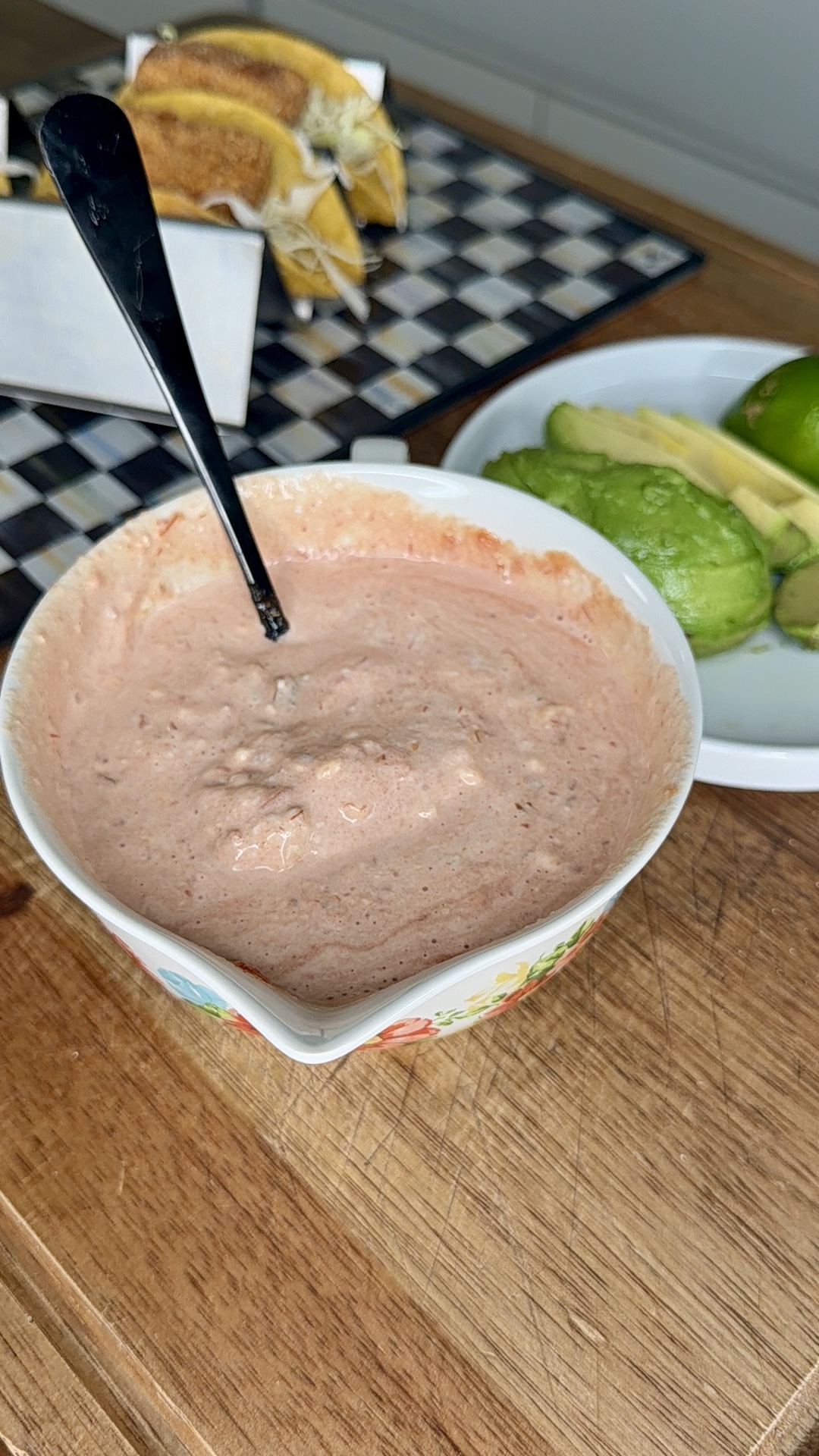 A bowl of creamy pink sauce with a spoon, sitting on a wooden surface. In the background, there’s a plate with sliced avocado and limes, and a tray of tacos.