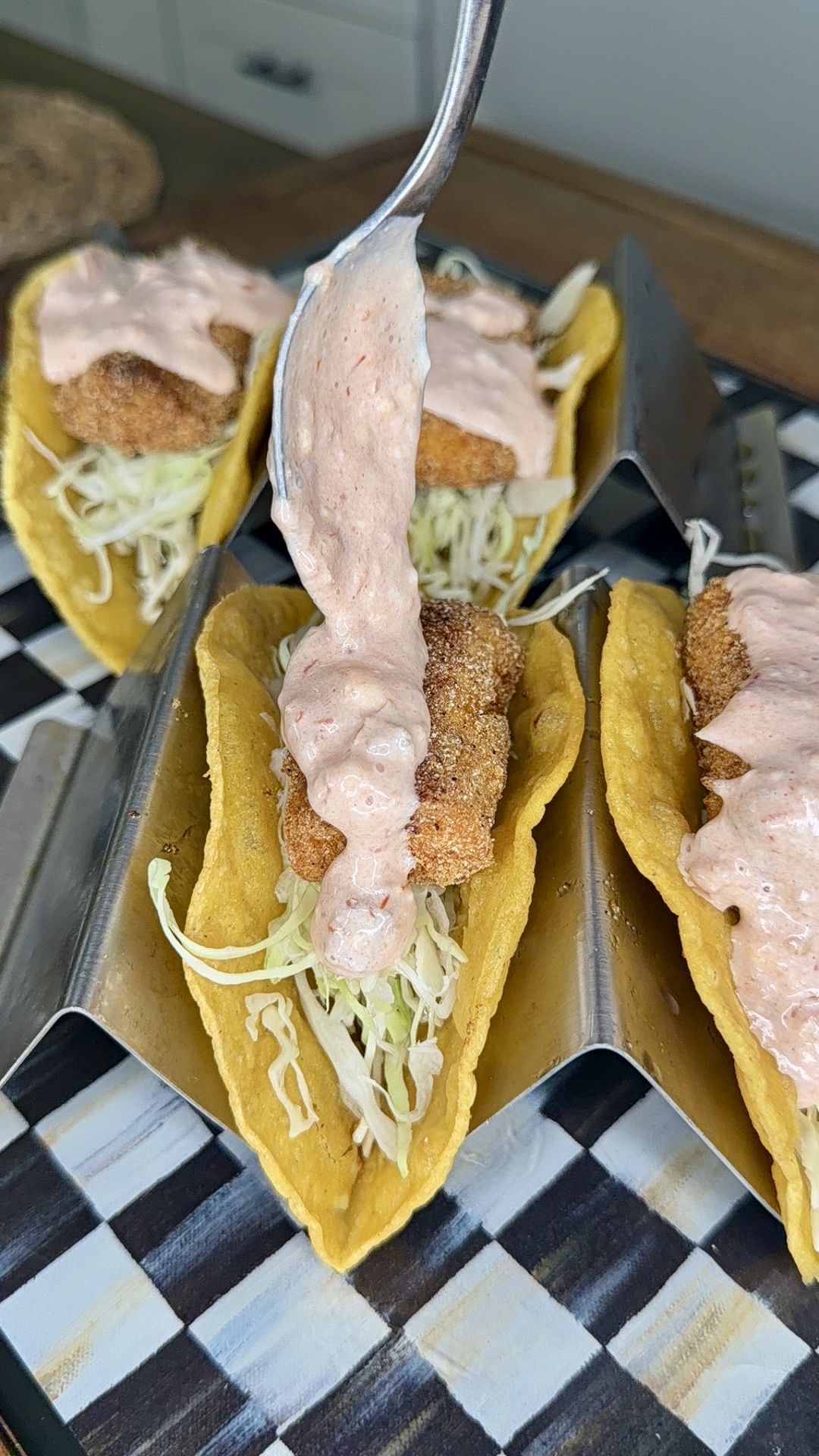 A spoon drizzles creamy sauce over crispy fish tacos filled with shredded lettuce in yellow corn tortillas, arranged in a metal taco holder on a checkered tray.