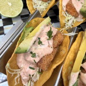 Three crispy fish tacos in taco holders, topped with shredded cabbage, sliced avocado, creamy pink sauce, and chopped herbs. A halved lime and part of an avocado are visible in the background.