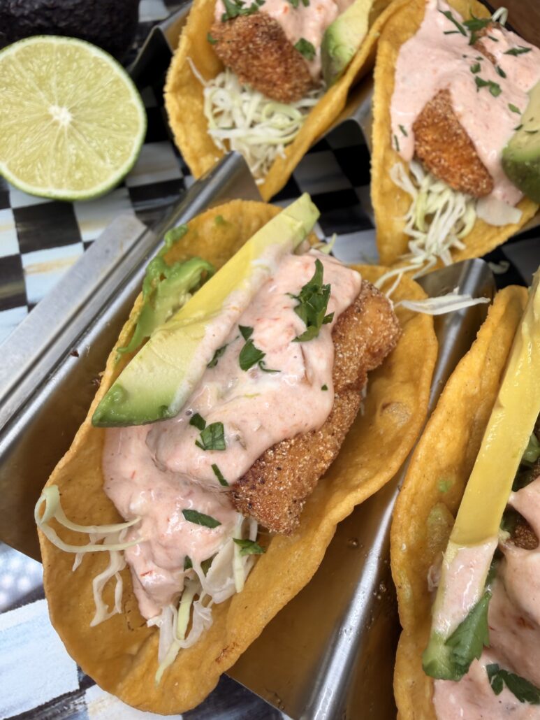 Three crispy fish tacos in taco holders, topped with shredded cabbage, sliced avocado, creamy pink sauce, and chopped herbs. A halved lime and part of an avocado are visible in the background.