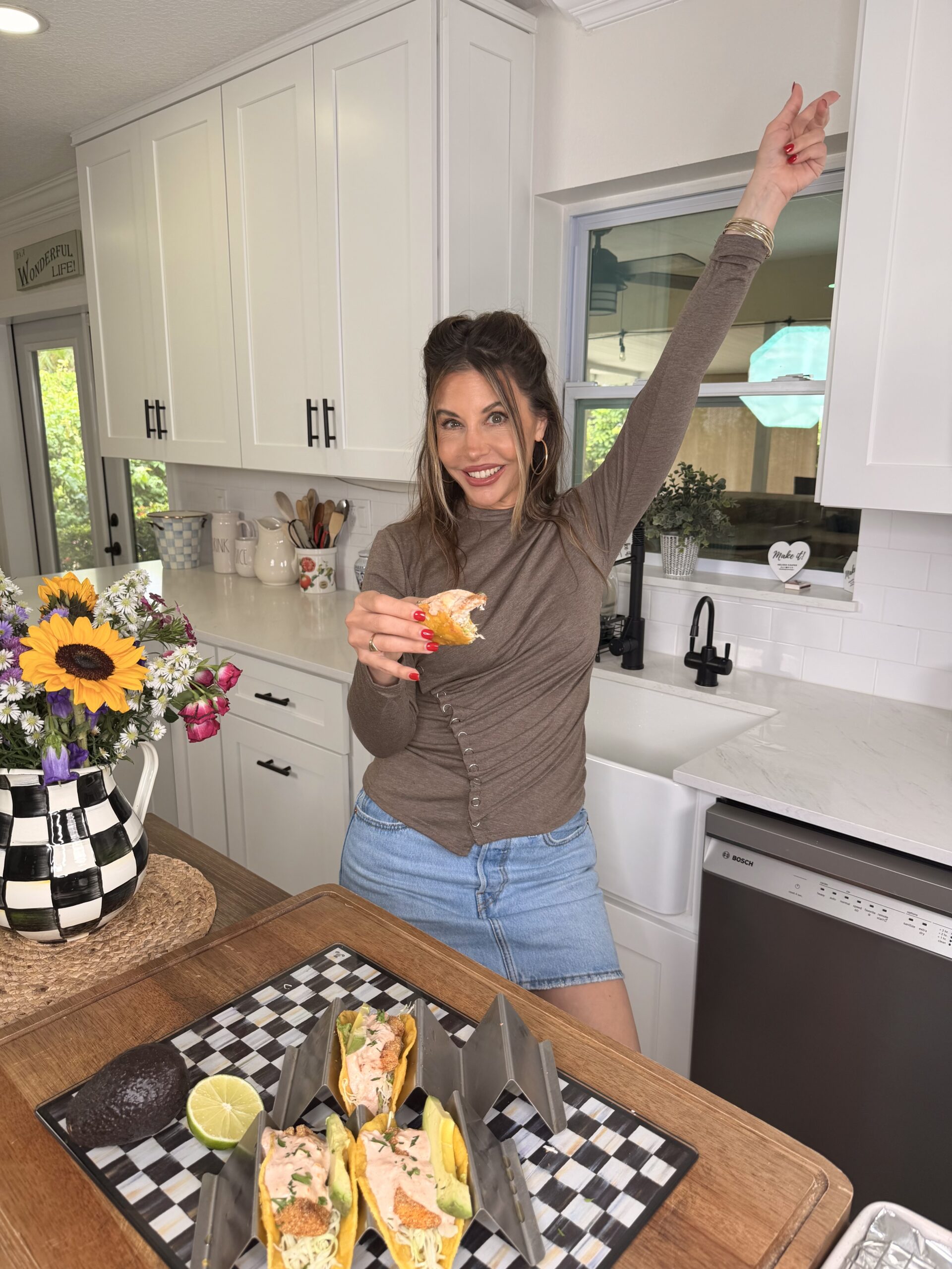 A woman in a brown top and denim skirt stands in a bright kitchen, smiling and pointing upward with one hand while holding a taco in the other. On the counter are tacos, a half avocado, a lime, and a vase of flowers.