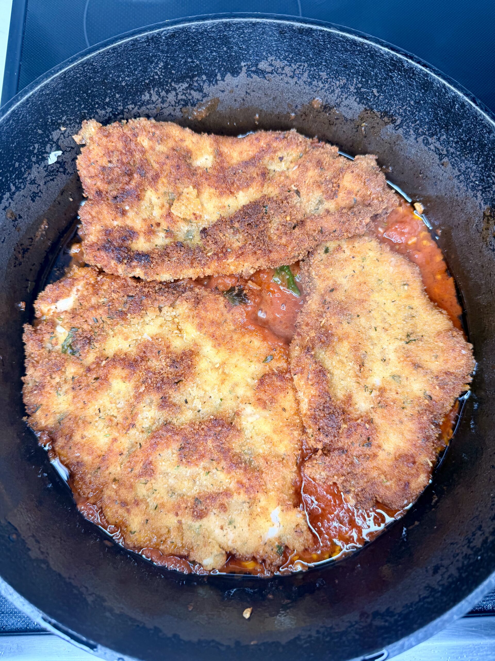 Three breaded and fried chicken cutlets are cooking in a black skillet, partially covering a layer of tomato sauce underneath. The cutlets are golden brown and crispy around the edges.