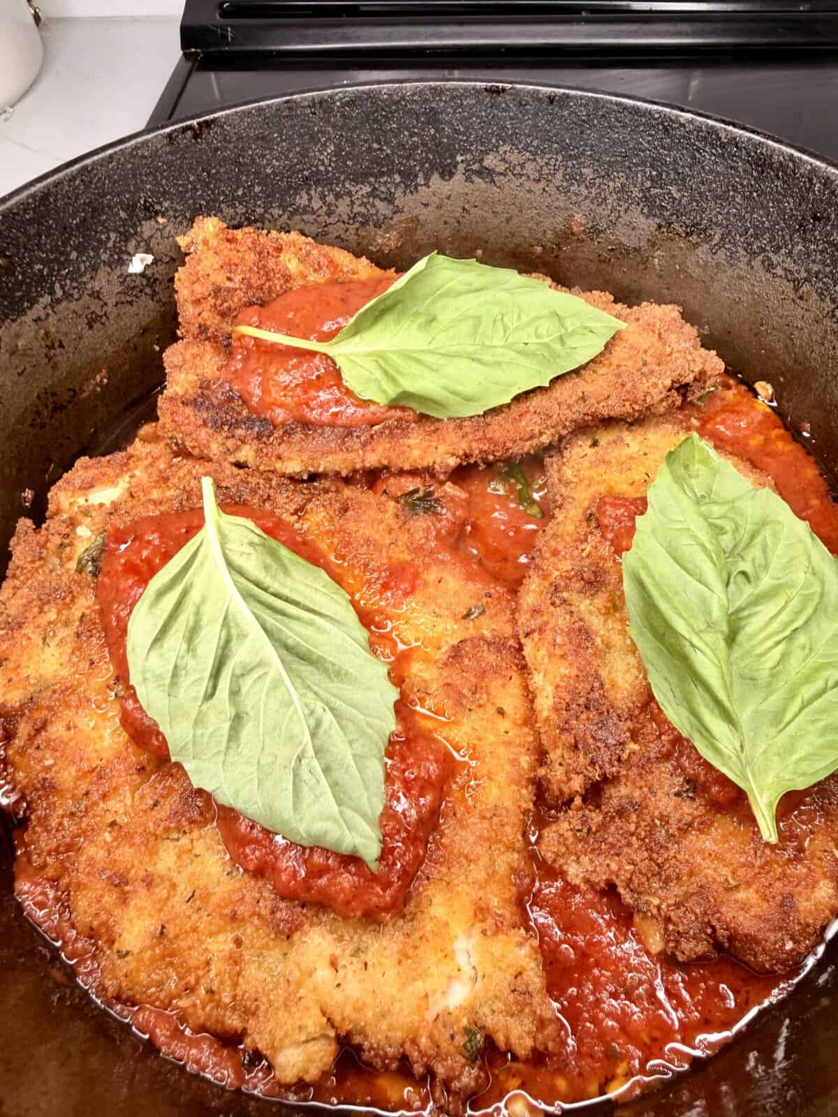 Three breaded and fried chicken cutlets topped with marinara sauce and fresh basil leaves are shown in a pan, ready to serve.