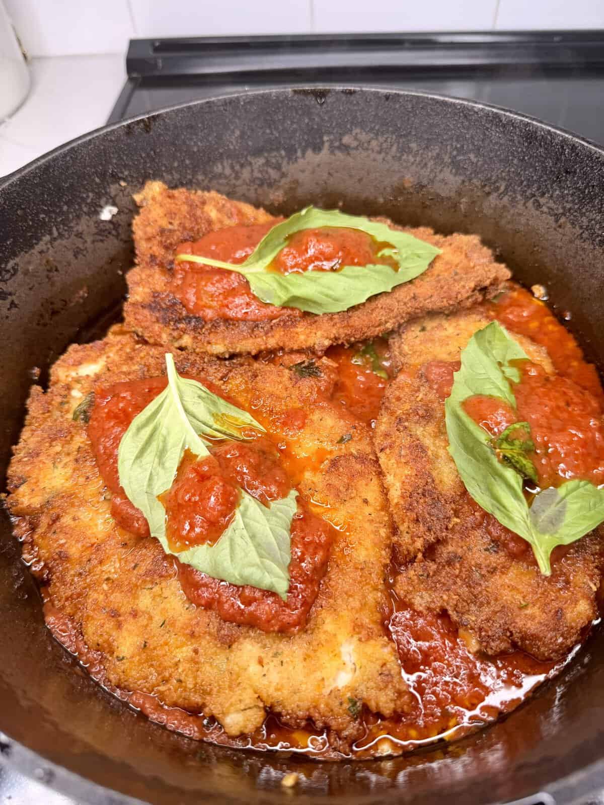 Breaded and fried chicken cutlets topped with tomato sauce and fresh basil leaves, arranged in a black cast iron skillet.