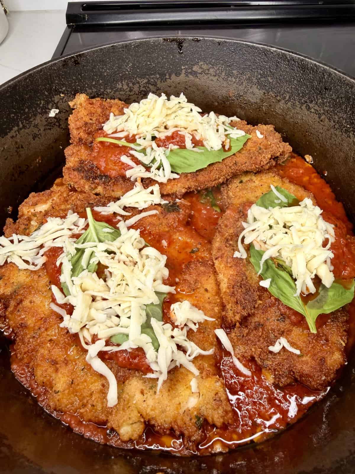 Breaded chicken cutlets in a cast iron pan topped with tomato sauce, fresh basil leaves, and shredded mozzarella cheese, ready to be baked or finished cooking for chicken parmesan.