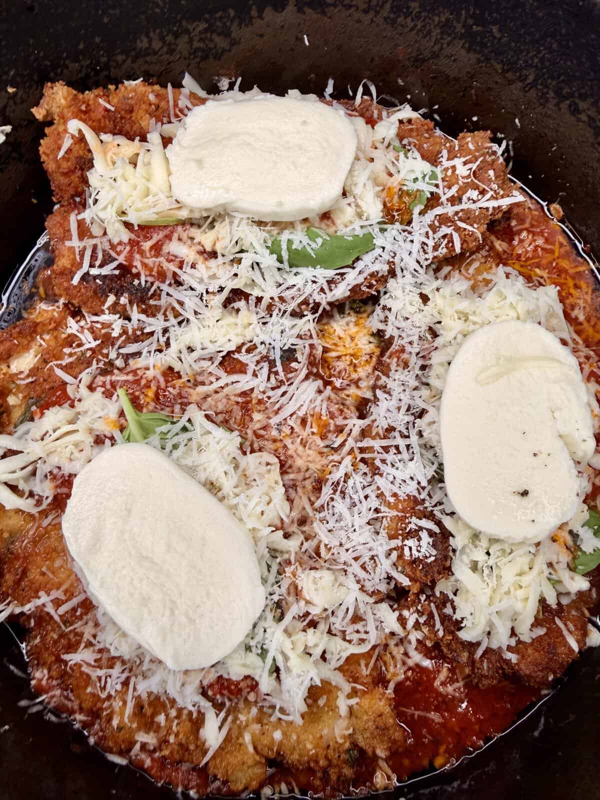 Breaded chicken cutlets topped with tomato sauce, shredded cheese, herbs, and three slices of fresh mozzarella, all in a pan, beginning to melt together for chicken parmesan.