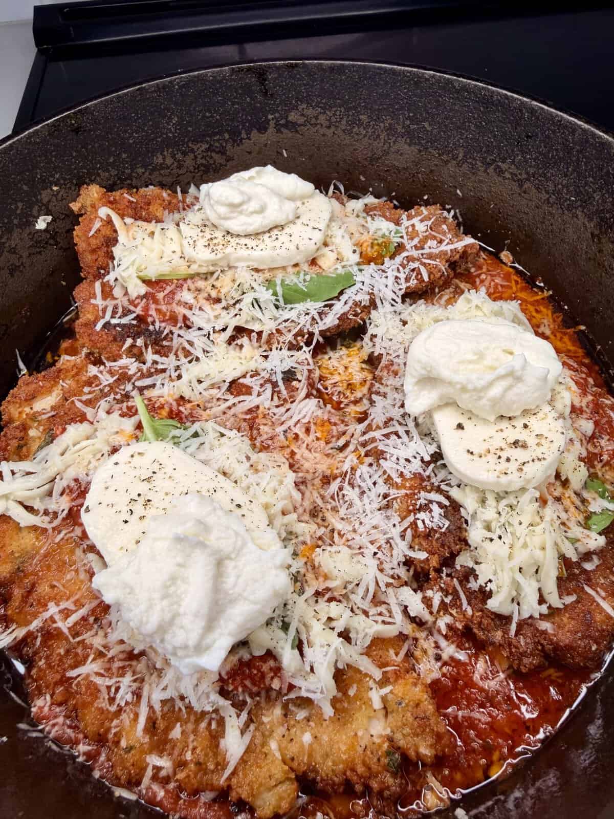 Breaded chicken cutlets topped with tomato sauce, shredded cheese, mozzarella slices, and dollops of ricotta cheese in a black cast iron pan, ready to be baked for chicken parmesan.