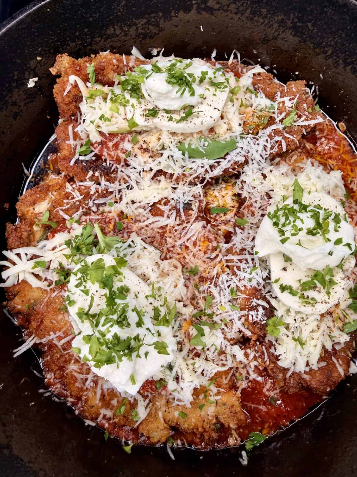 A close-up of chicken parmesan in a skillet, topped with tomato sauce, melted cheese, fresh mozzarella, chopped parsley, and grated parmesan. The dish has a crispy, golden-brown crust and is garnished with herbs.