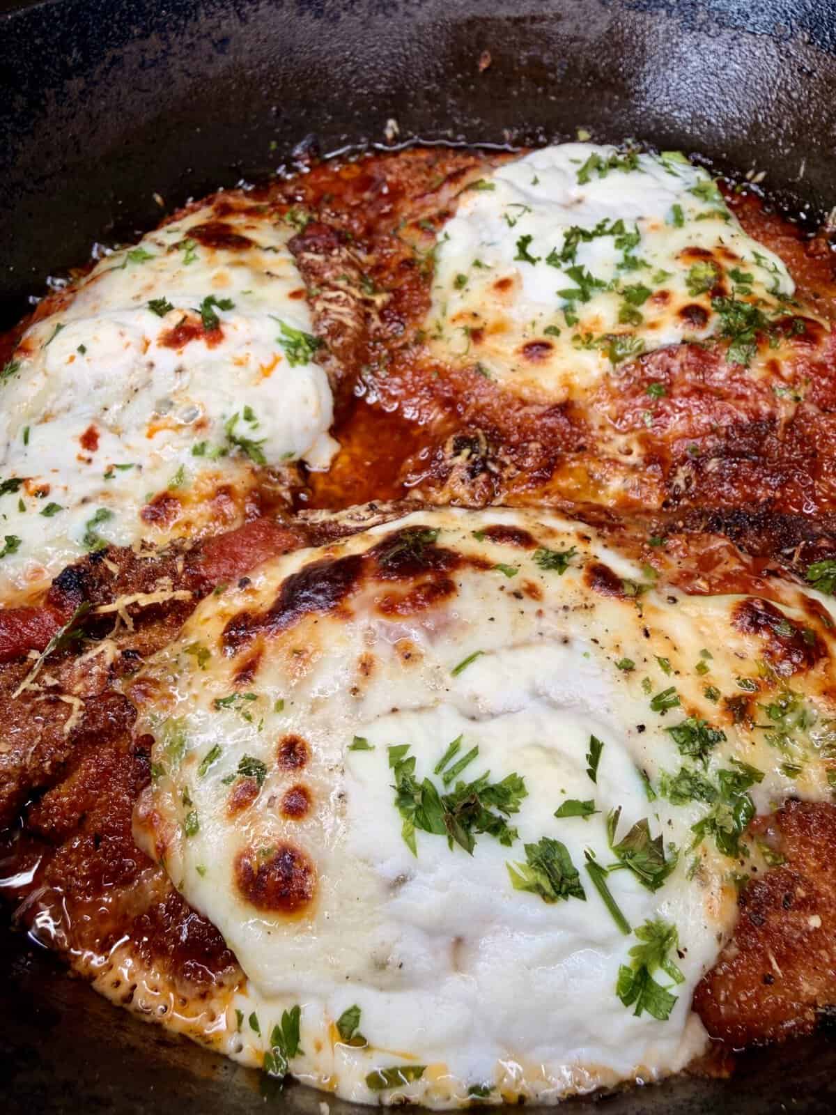 Close-up of chicken parmesan in a skillet, topped with melted mozzarella cheese, tomato sauce, and sprinkled with fresh parsley. The edges of the cheese and sauce are golden and slightly crispy.
