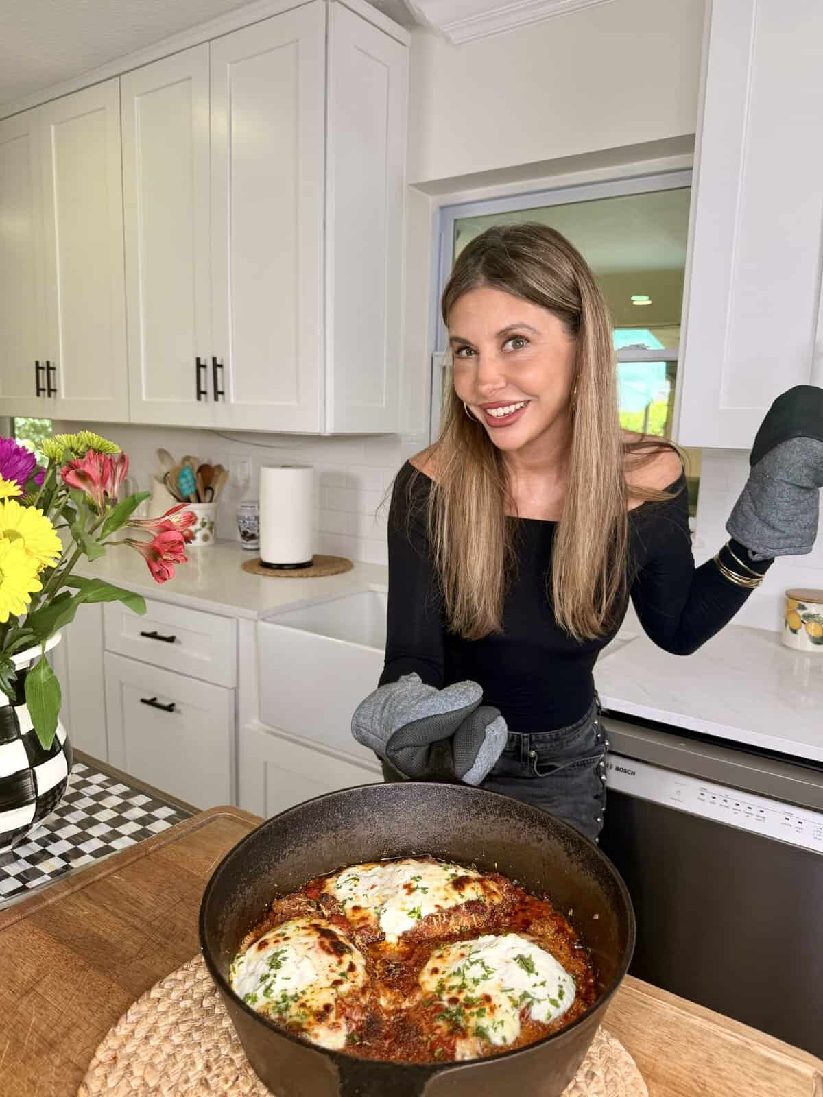 A smiling woman in a black top and oven mitts stands in a bright kitchen, holding a skillet with baked eggs in tomato sauce, garnished with herbs. A bouquet of colorful flowers is on the counter beside her.