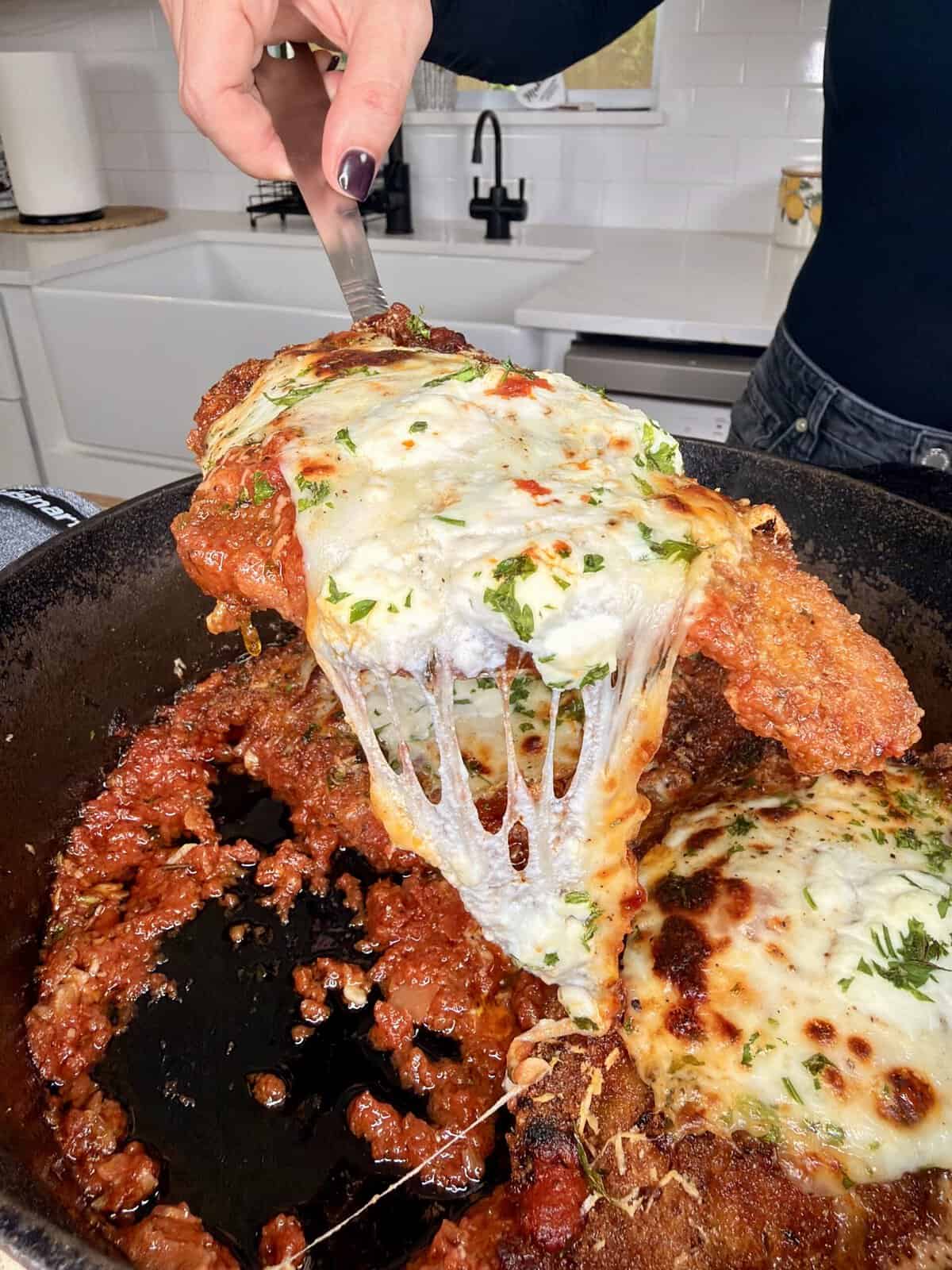 A hand lifts a slice of cheesy, baked chicken parmesan from a skillet, showing melted cheese stretching with rich tomato sauce and herbs on top. A white kitchen is visible in the background.