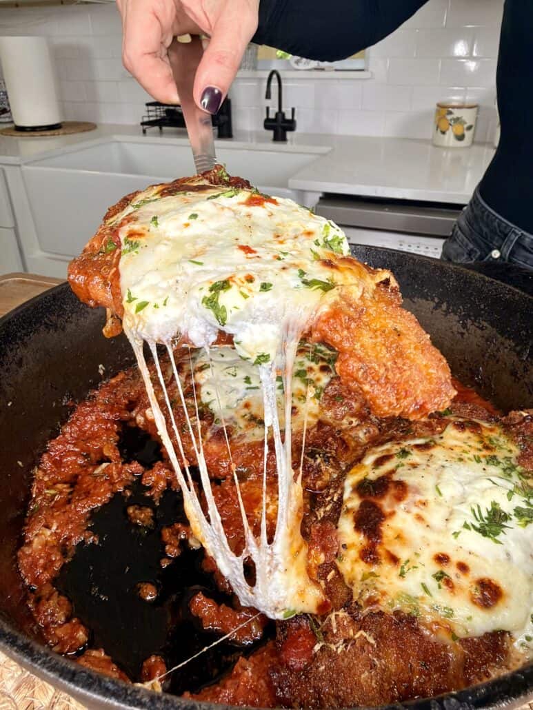 A hand lifts a cheesy, breaded chicken cutlet covered in marinara sauce and melted mozzarella from a cast iron skillet, with gooey cheese stretching between the skillet and the cutlet. The kitchen is visible in the background.