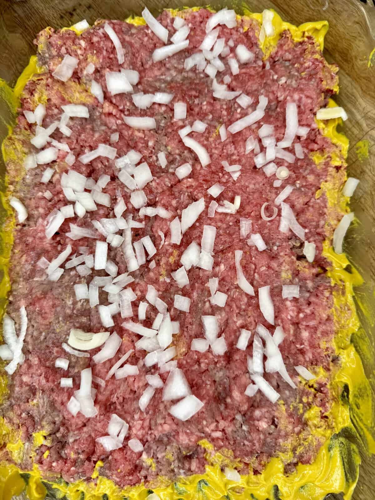 A baking dish filled with raw ground beef spread over a layer of yellow mustard, topped with chopped white onions.