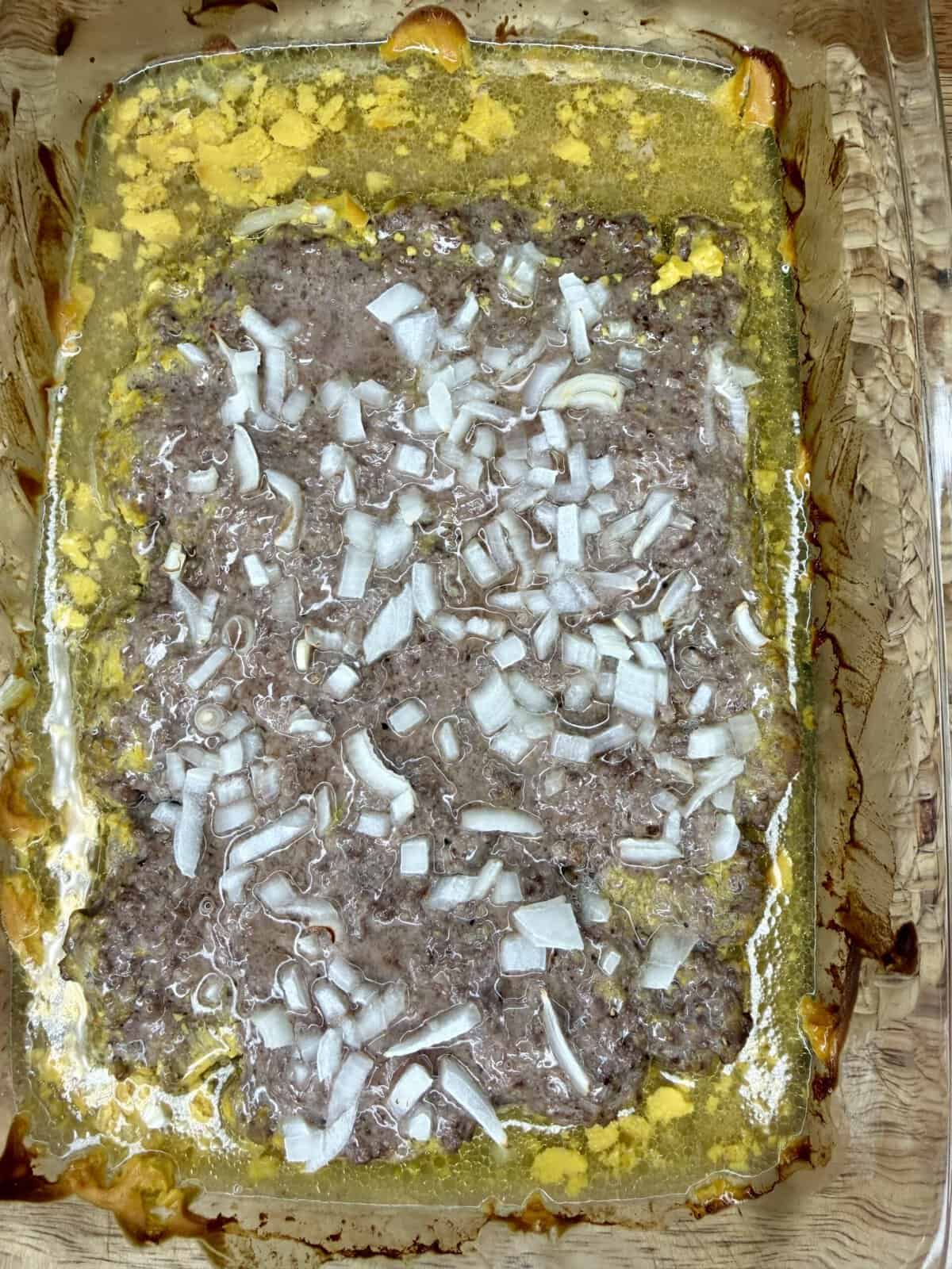 A glass baking dish filled with ground meat topped with chopped onions, surrounded by yellowish melted fat and juices, with browned edges around the sides of the dish.