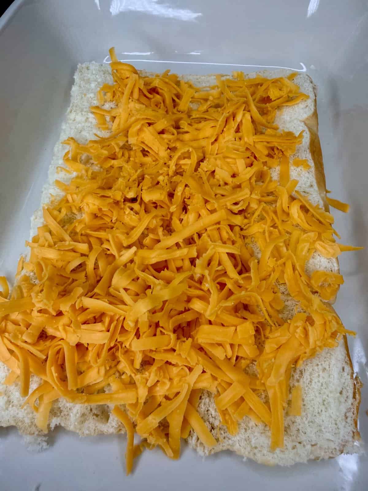 Shredded cheddar cheese spread evenly over a layer of white bread slices in a white baking dish.