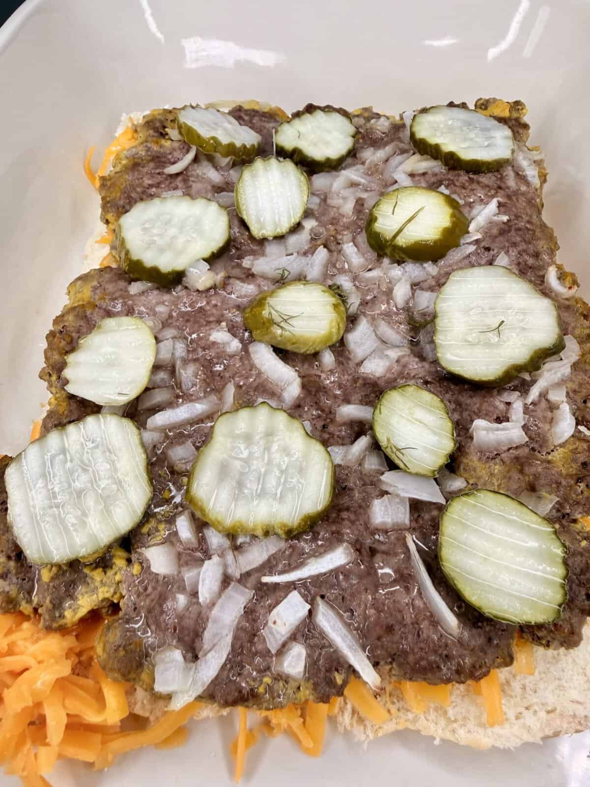 A close-up of an uncooked layered burger casserole with shredded cheese, ground beef, chopped onions, and sliced pickles on a layer of bread in a baking dish.