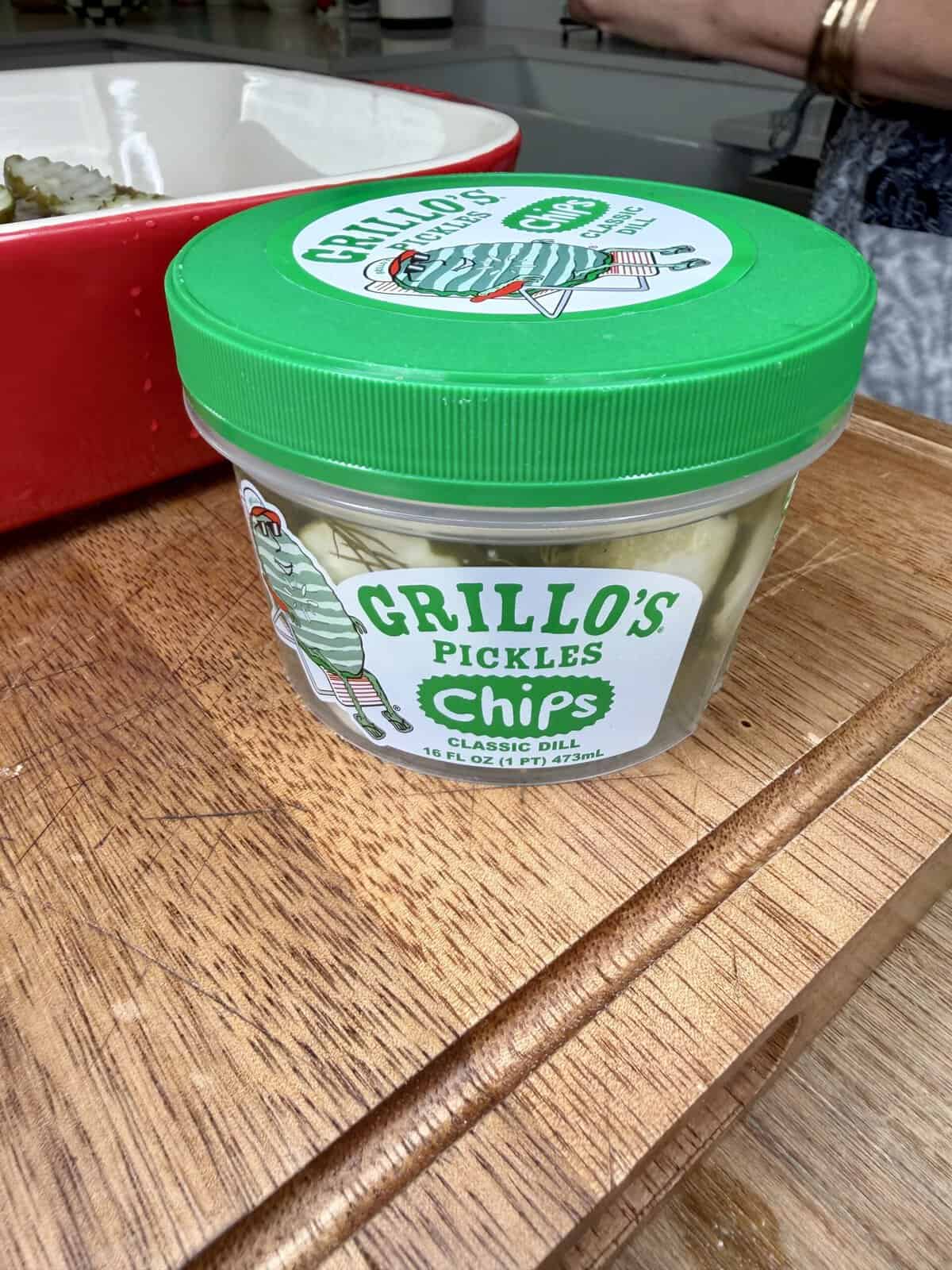 A container of Grillo’s Pickles Chips, Classic Dill flavor, sits on a wooden cutting board. Part of a red tray and a person’s arm are visible in the background.