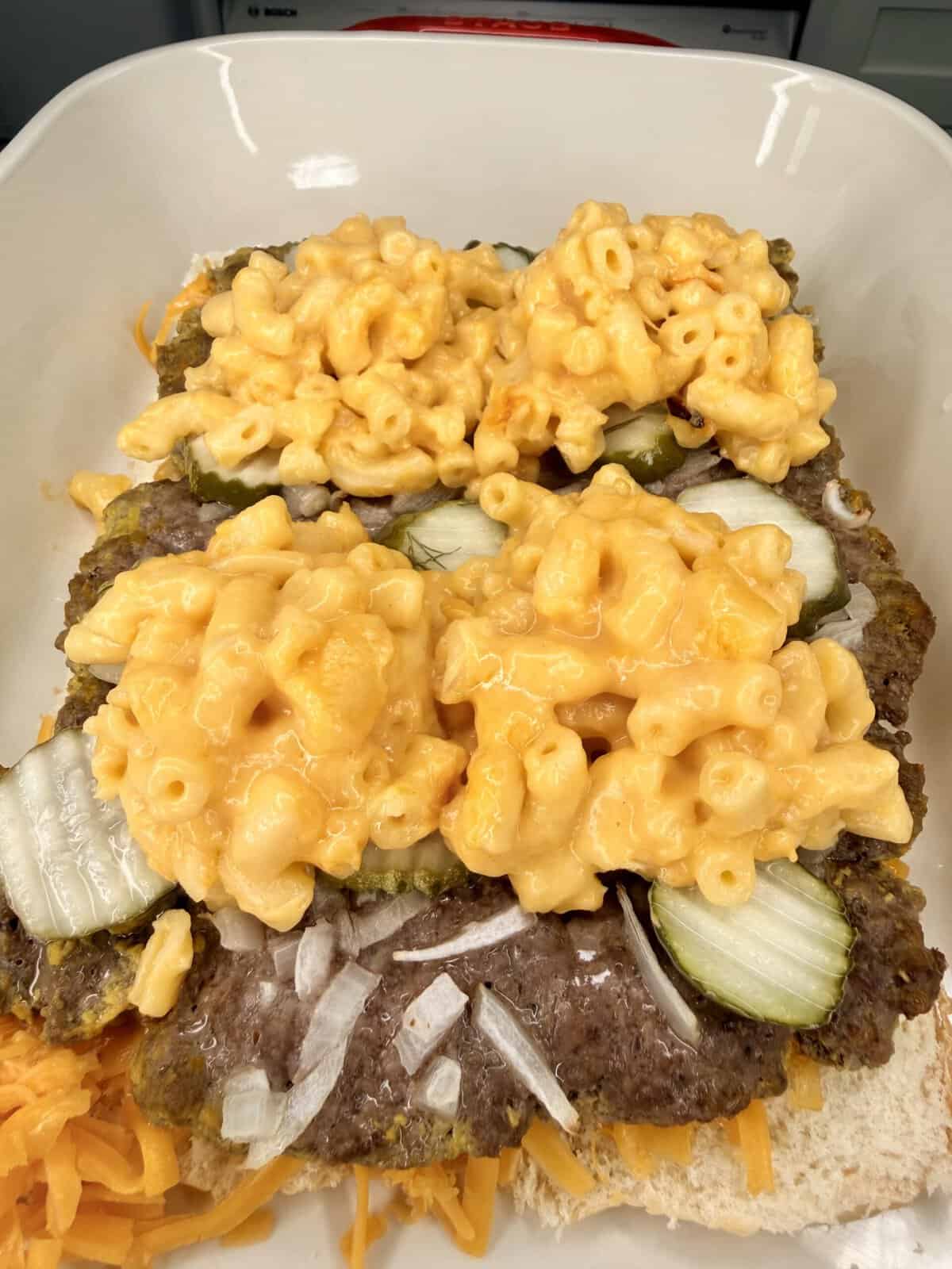 A close-up of a sandwich topped with hamburger patties, sliced pickles, chopped onions, shredded cheddar cheese, and generous scoops of macaroni and cheese on a white dish.