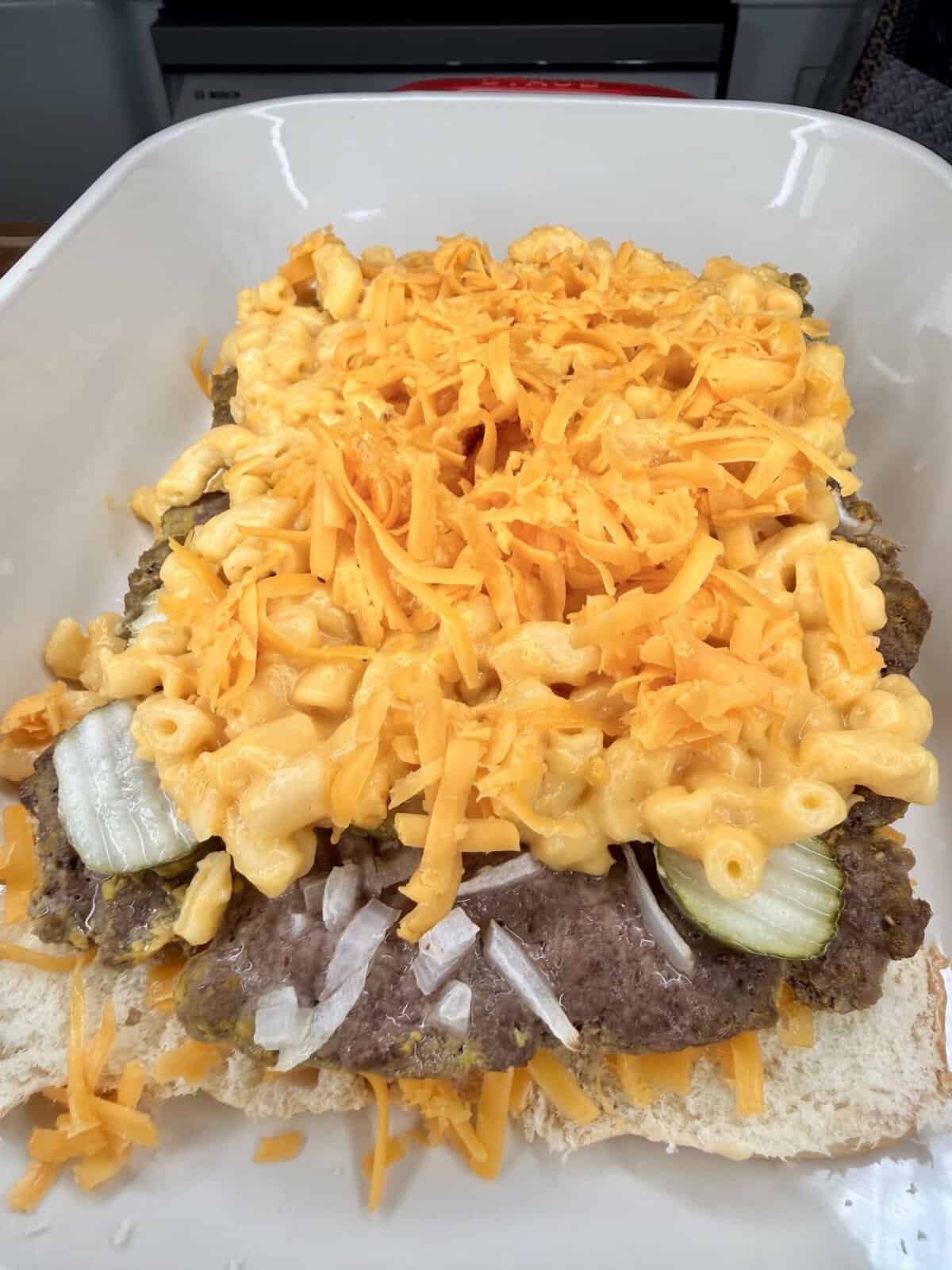 A close-up of an open sandwich bun topped with hamburger patties, pickles, diced onions, macaroni and cheese, and a generous amount of shredded cheddar cheese.