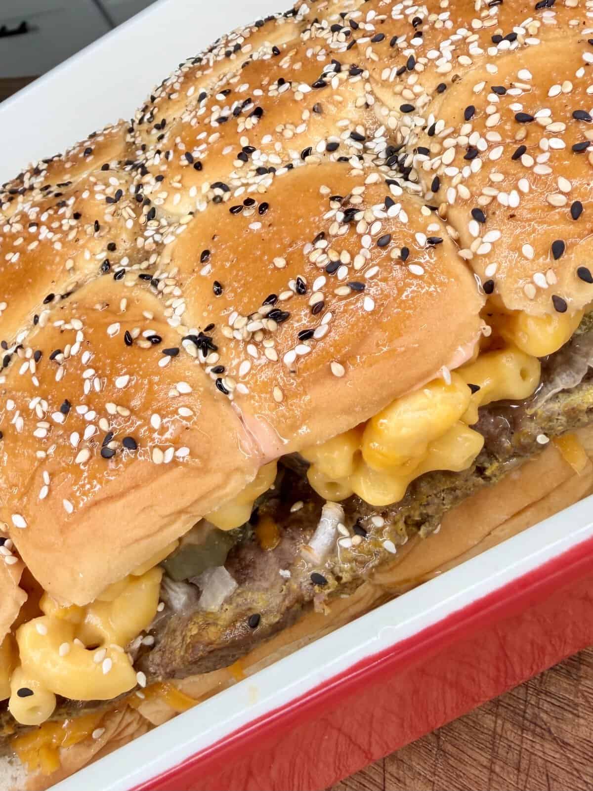 Close-up of baked sandwiches in a red dish, featuring sesame-seeded buns filled with creamy macaroni and cheese and what appears to be ground beef patties or meatloaf.