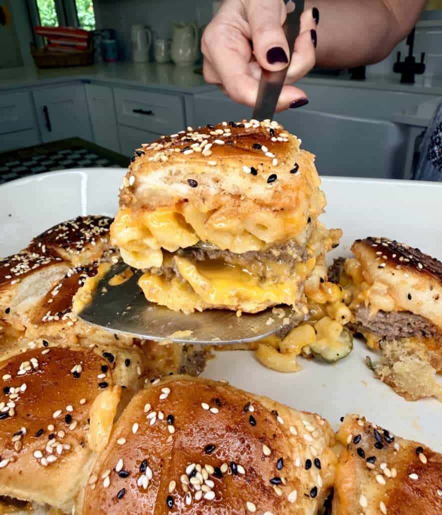A hand lifts a cheesy macaroni and beef slider topped with sesame seeds from a tray of similar sliders in a bright kitchen.