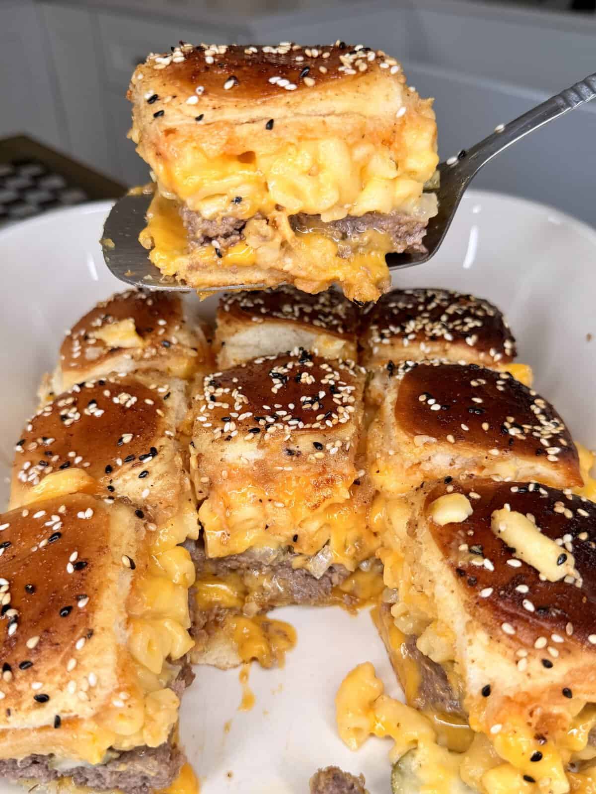 A close-up of cheesy sliders topped with sesame seed buns, filled with macaroni and cheese and meat, with one slider being lifted by a spatula above a white dish containing the rest.