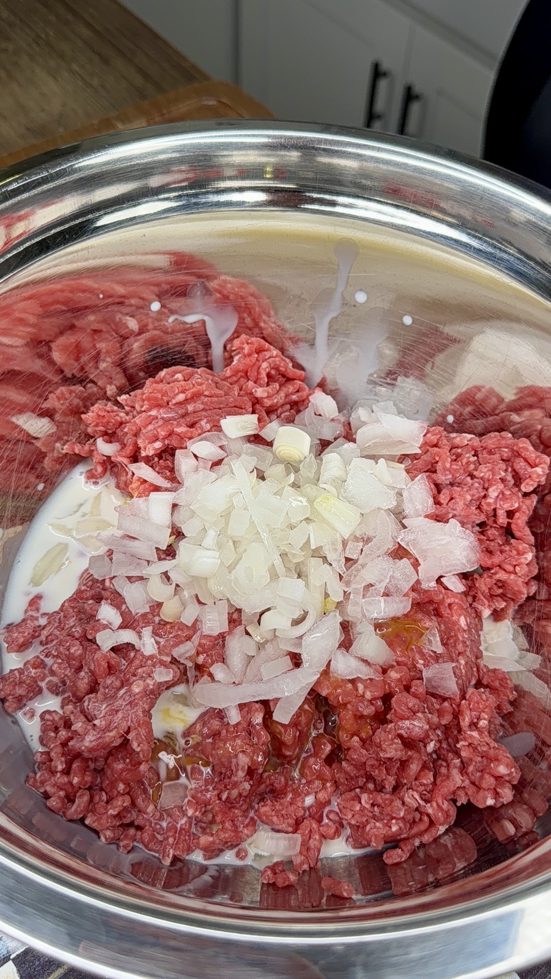 A metal bowl containing ground beef, chopped onions, a splash of milk, and a cracked egg, ready to be mixed for a recipe.
