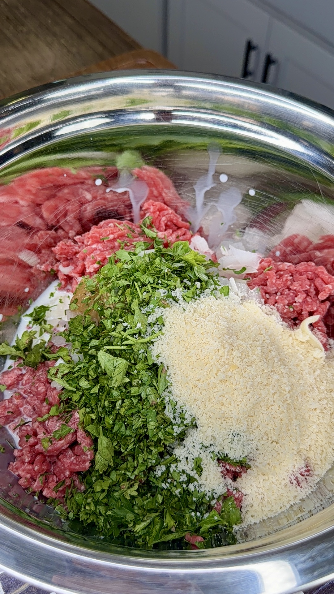 A metal bowl filled with raw ground meat, chopped fresh herbs, grated cheese, and other ingredients, ready to be mixed. Kitchen cabinets and a countertop are visible in the background.
