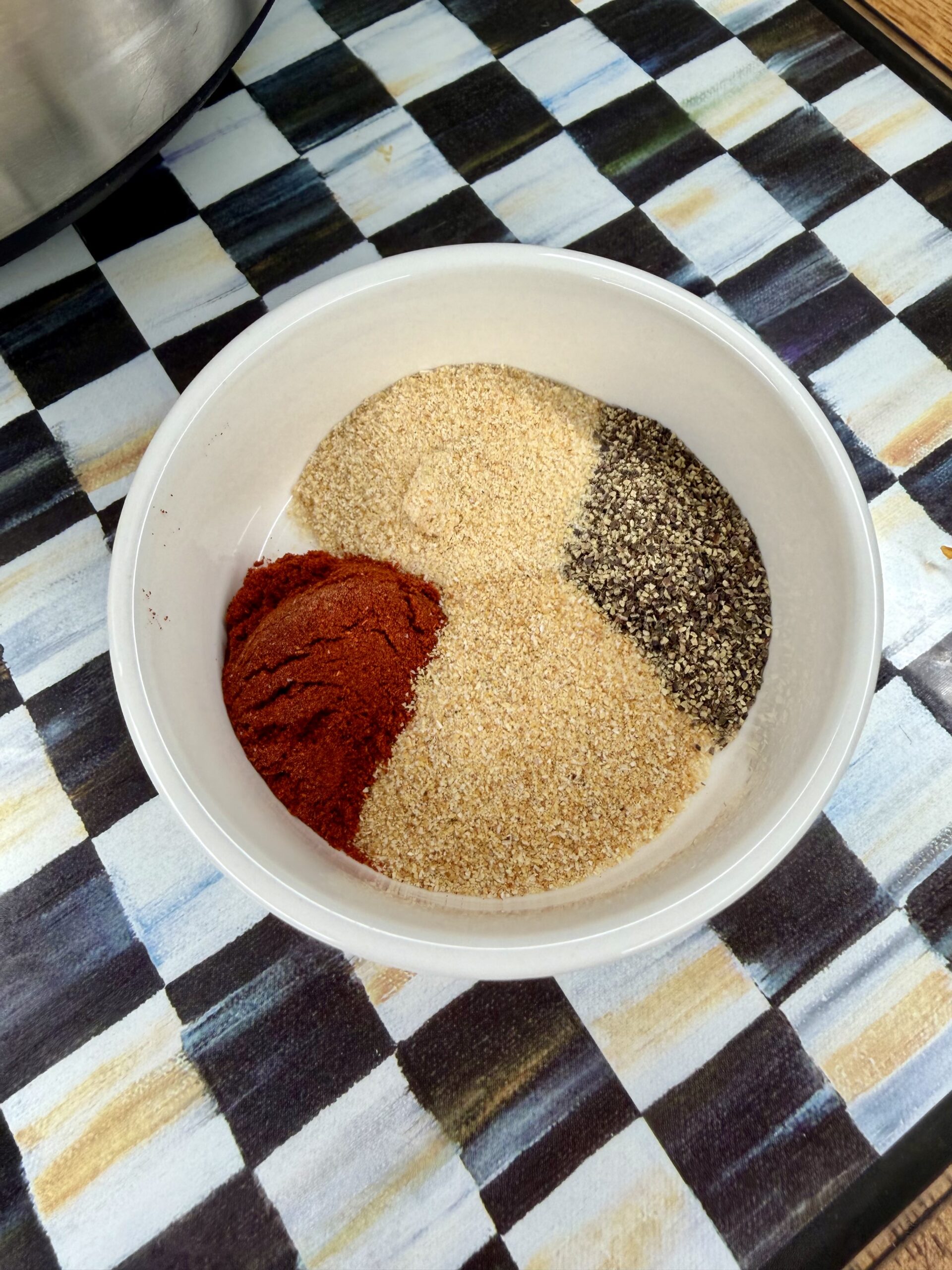 A white bowl containing four piles of seasonings: ground black pepper, paprika, onion powder, and garlic powder, placed on a checkered tablecloth.