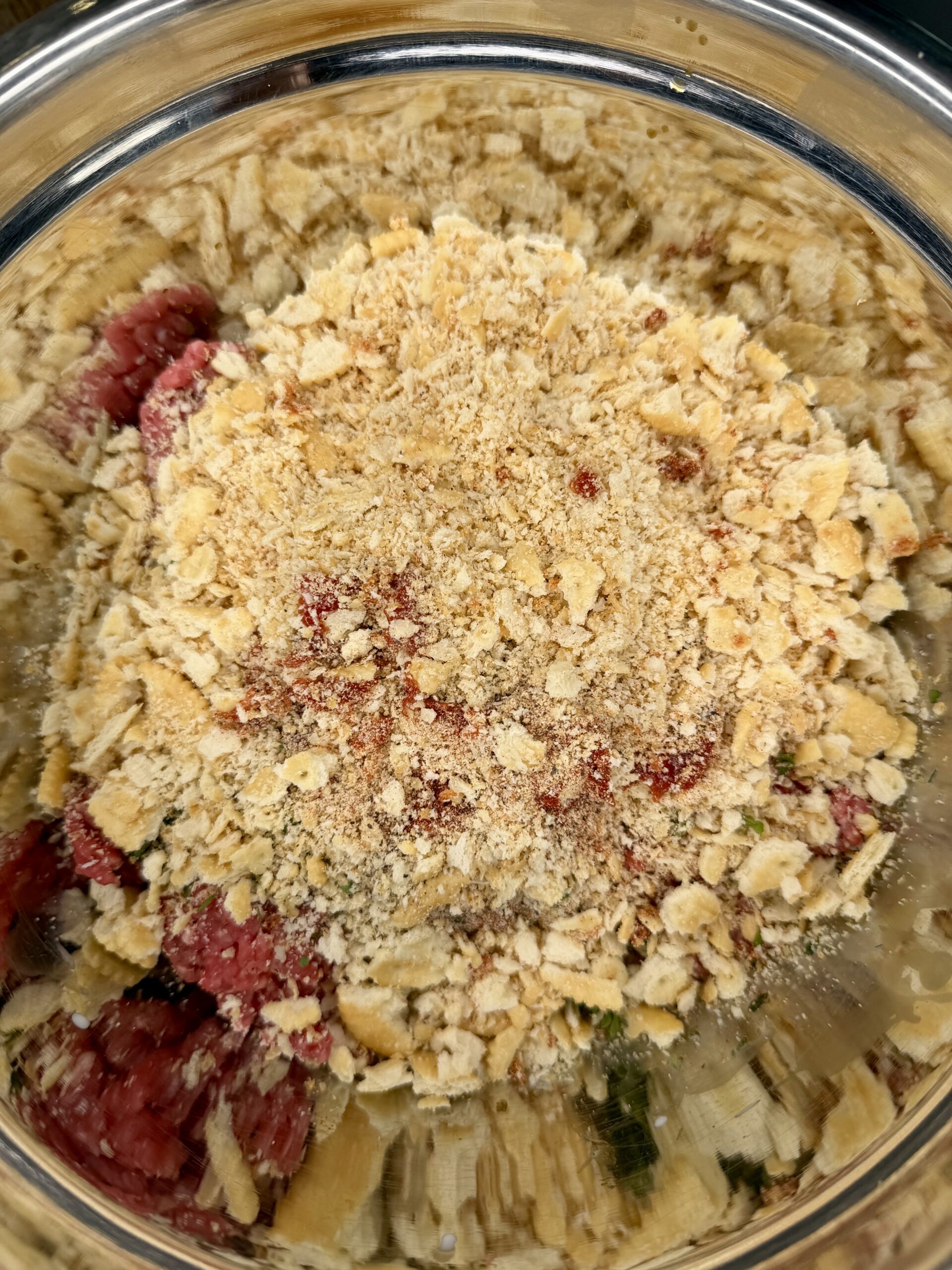 A metal bowl filled with ground meat and topped with a layer of crushed crackers, ready to be mixed for a recipe.