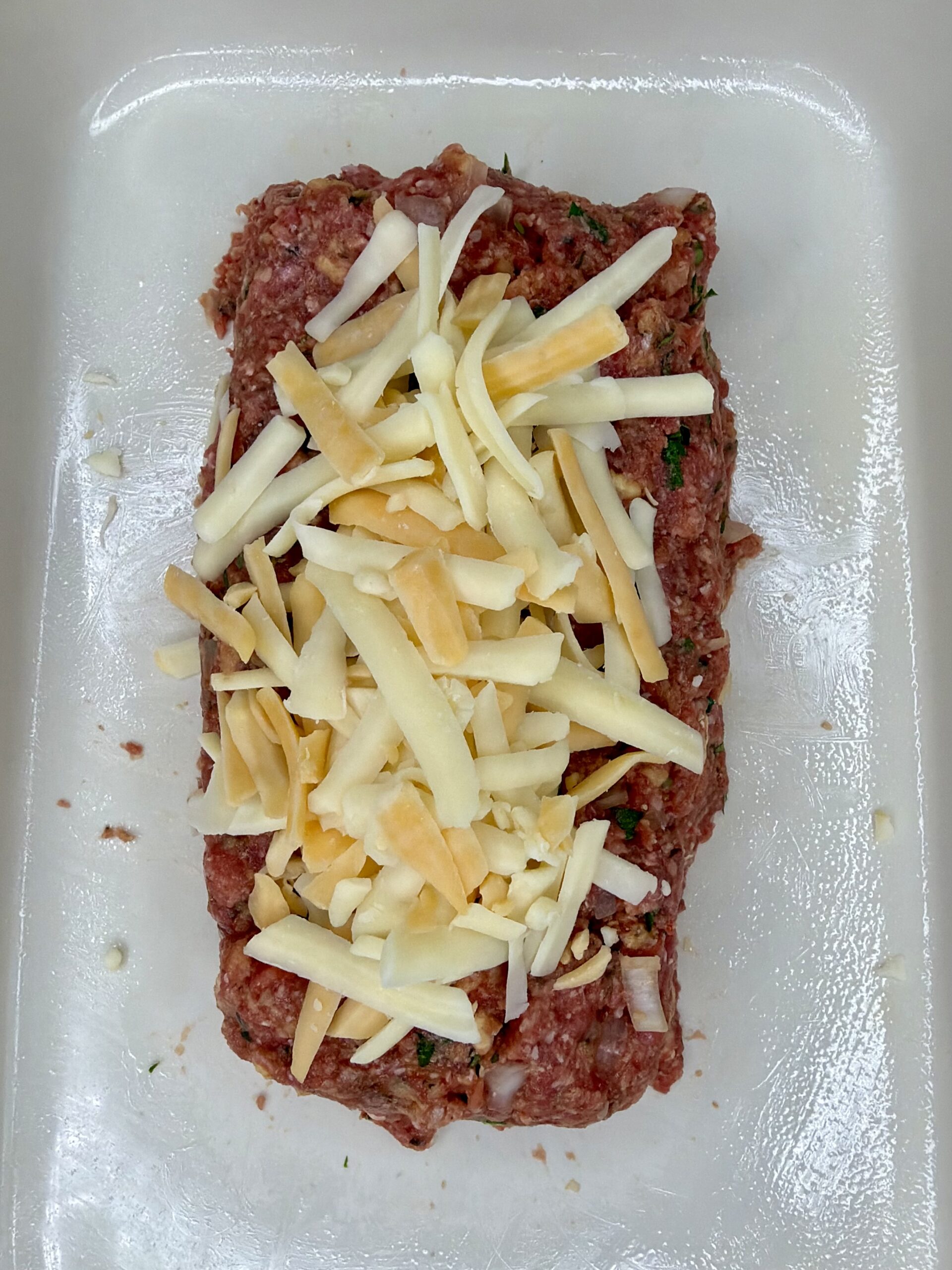 A raw meatloaf mixture shaped into a loaf sits on a glass tray, topped with a generous layer of shredded cheese, ready to be cooked.